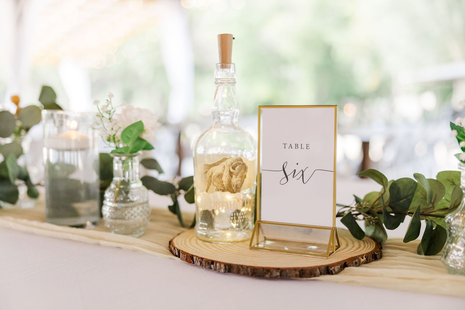 Elegant wedding table six centerpiece with a decorative bottle, gold-framed sign, candles, and greenery on a wood slice.