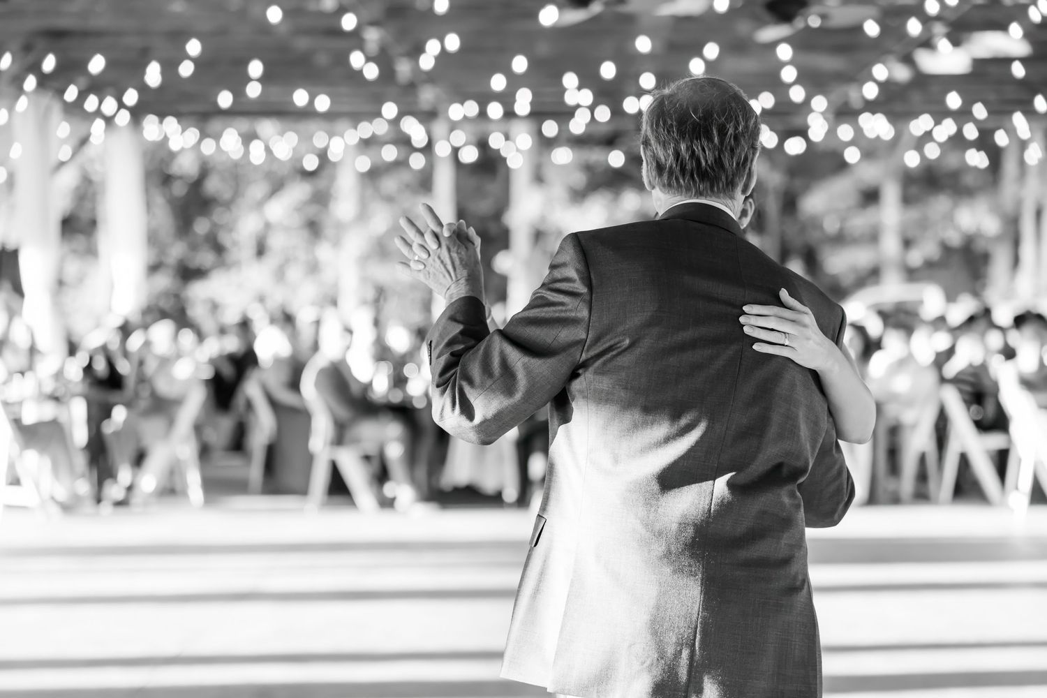 Black and white photo of bride and groom sharing first dance at wedding reception with string lights.