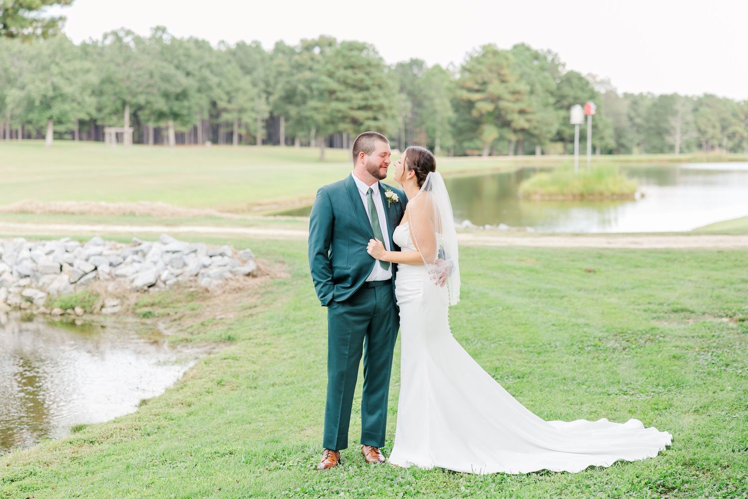 Newlyweds embrace on a lush green lawn with a serene pond and trees in the background.