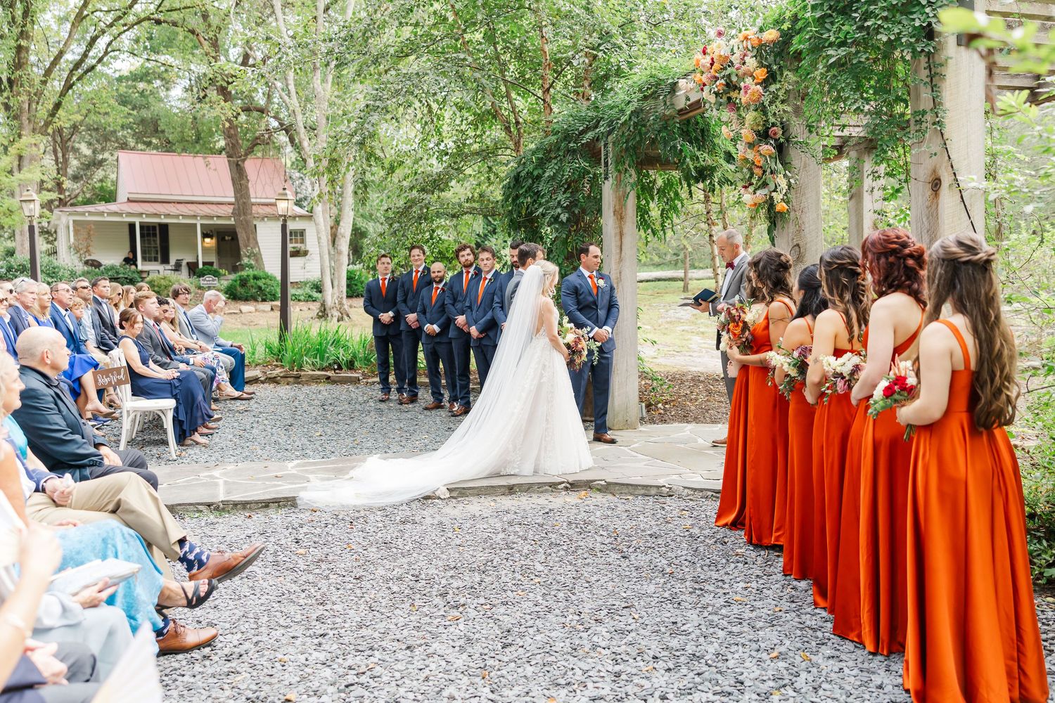 Wedding party stands in a row at outdoor ceremony with bridesmaids wearing vibrant burnt orange dresses.