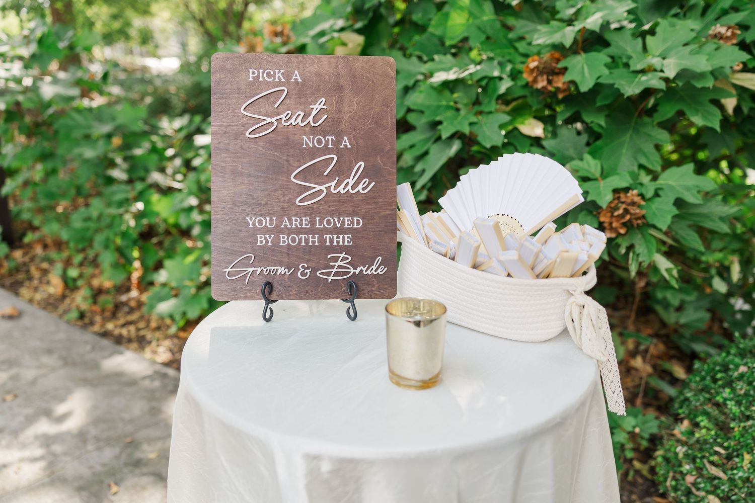 A wooden wedding sign sits on a table with a white tablecloth and floral centerpiece in an outdoor garden setting.