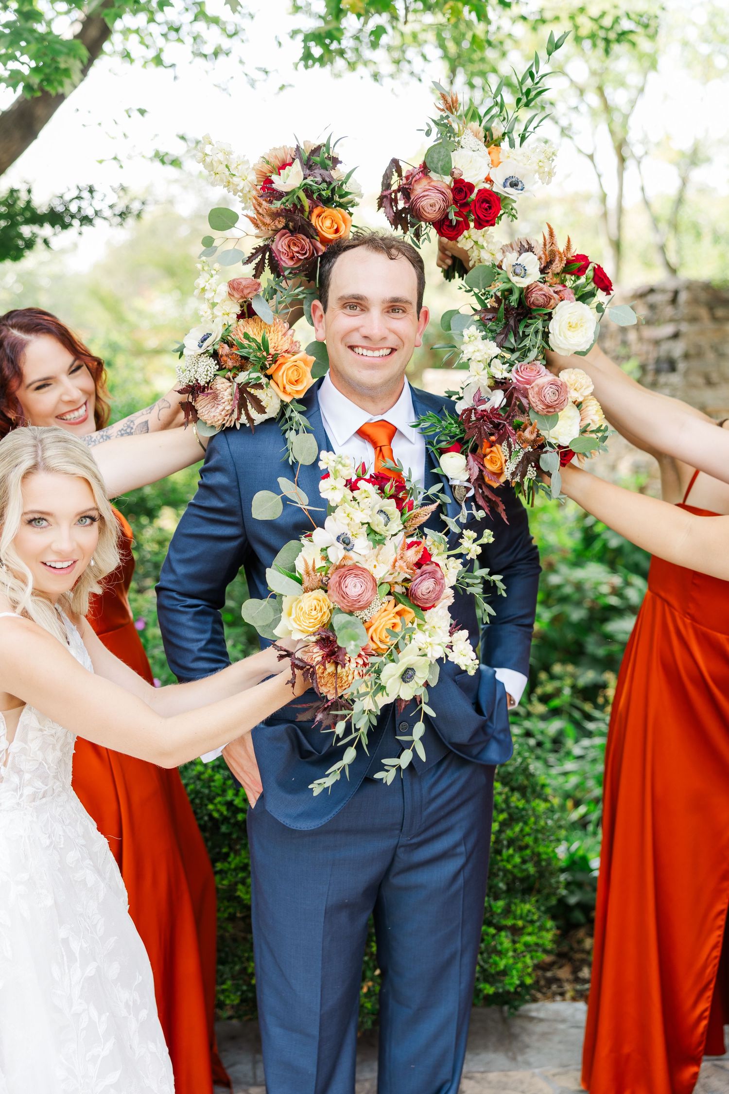 Wedding party in navy suits and burnt orange dresses pose with vibrant floral bouquets in an outdoor garden setting.
