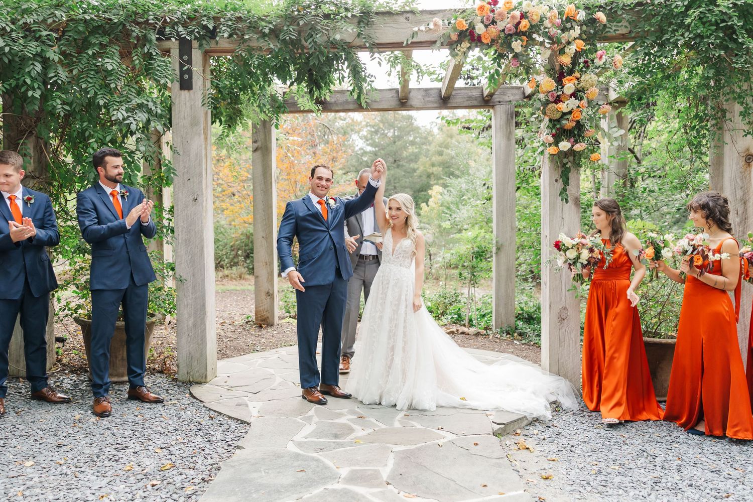 A wedding party poses in an outdoor garden setting with bridesmaids wearing vibrant orange dresses.