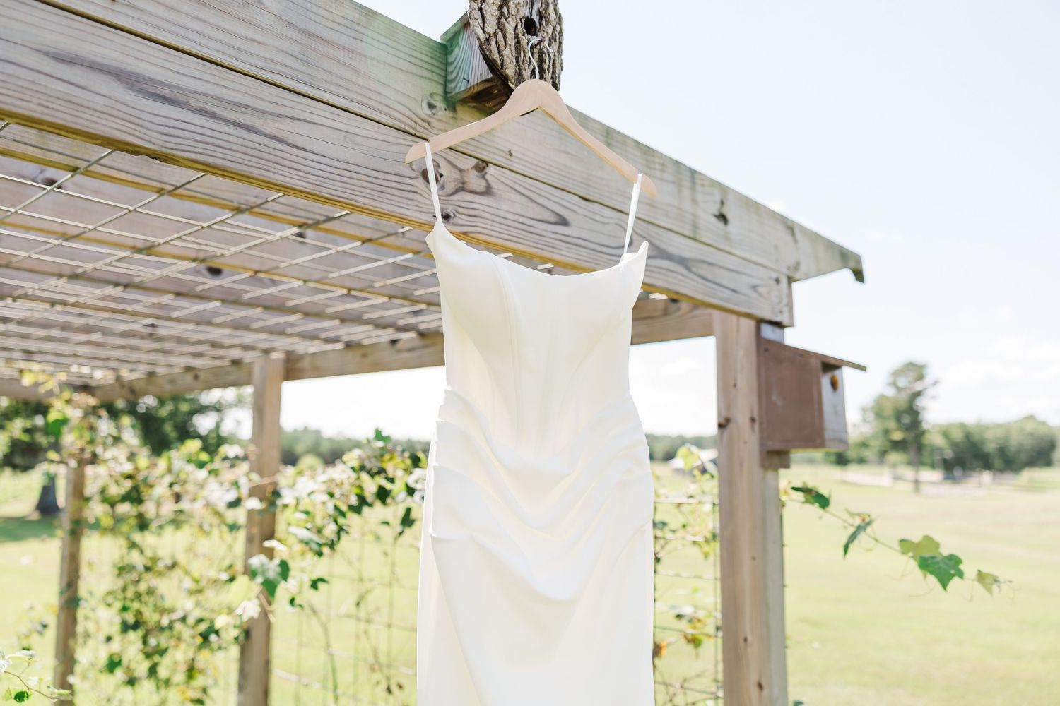 A white dress suspended from a wooden arbor structure with sunlit grass and trees in the background.
