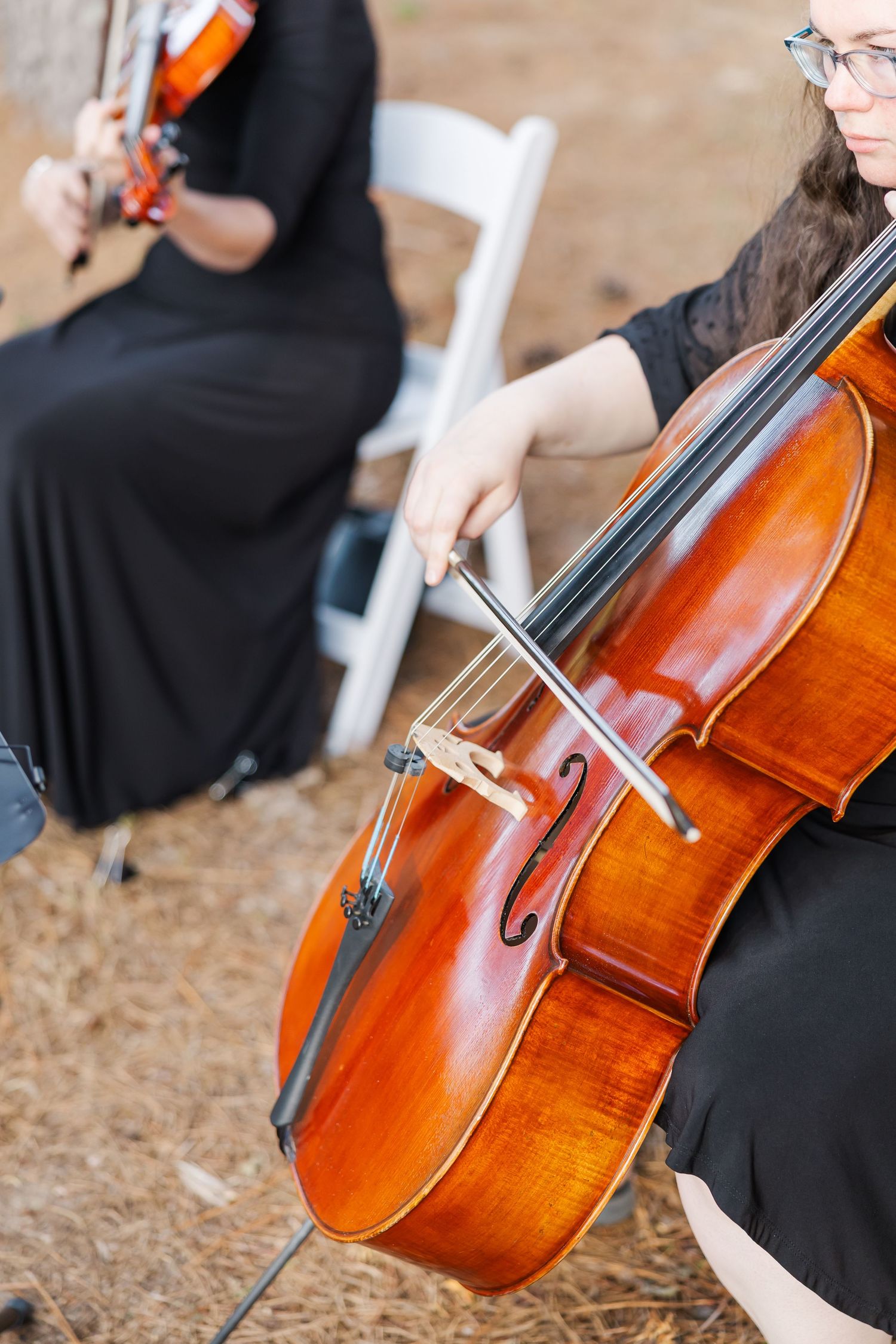 A musician in black plays cello with bow during an outdoor wedding ceremony alongside a violinist.