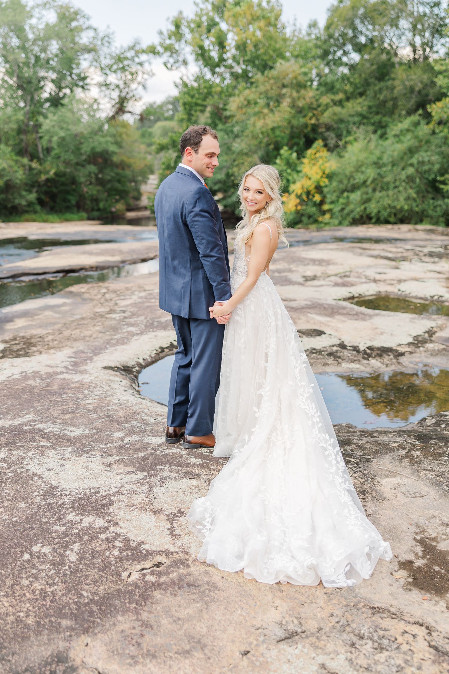A wedding couple stands on natural rock formations, with the bride's lace train flowing behind them.