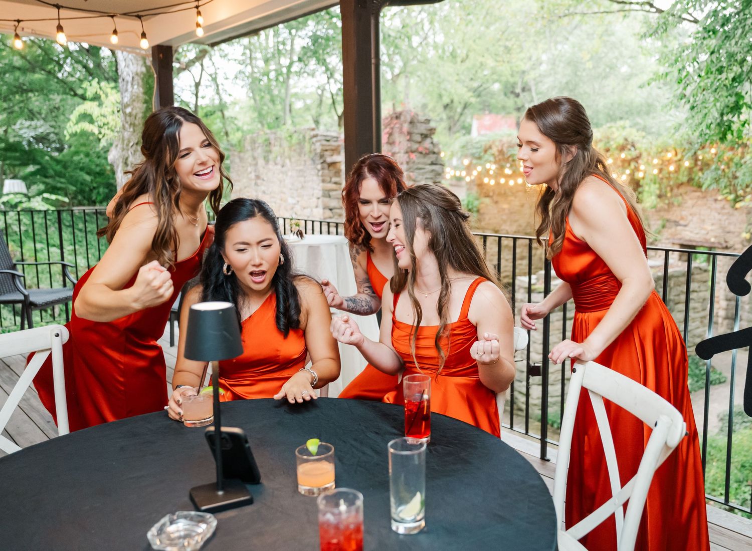 A group in orange and red dresses laughs together while seated at outdoor tables with string lights overhead.