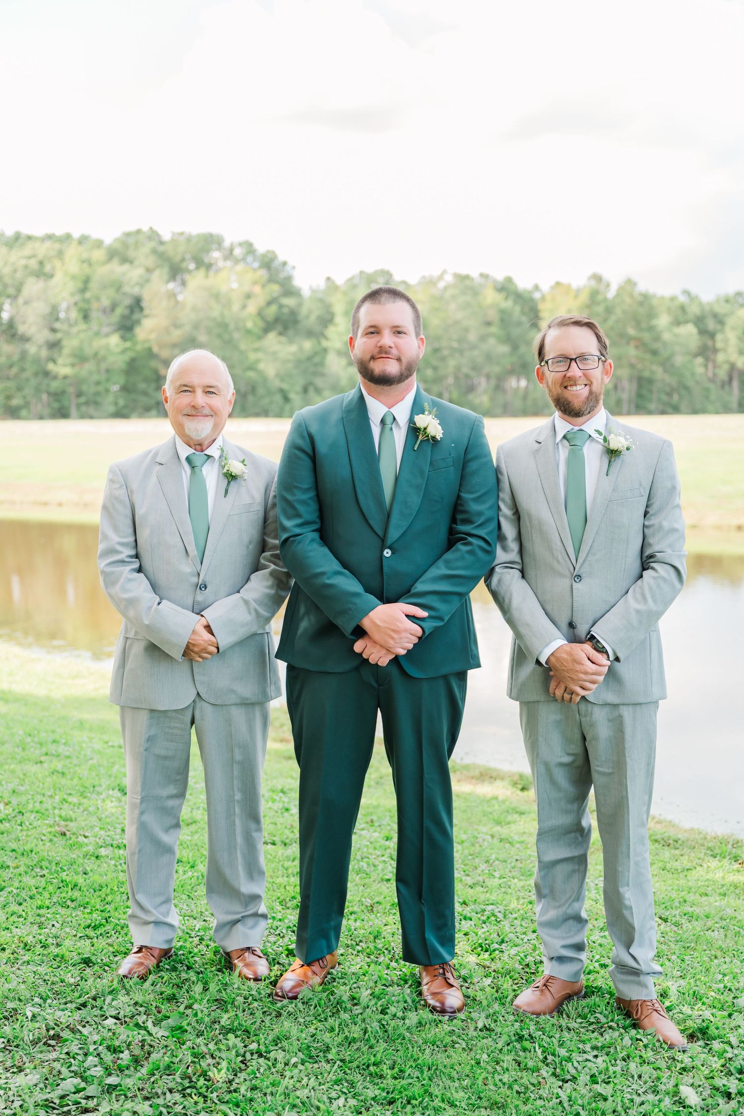 Groomsmen in light gray suits and one in emerald green pose together on grassy lawn by waterfront.