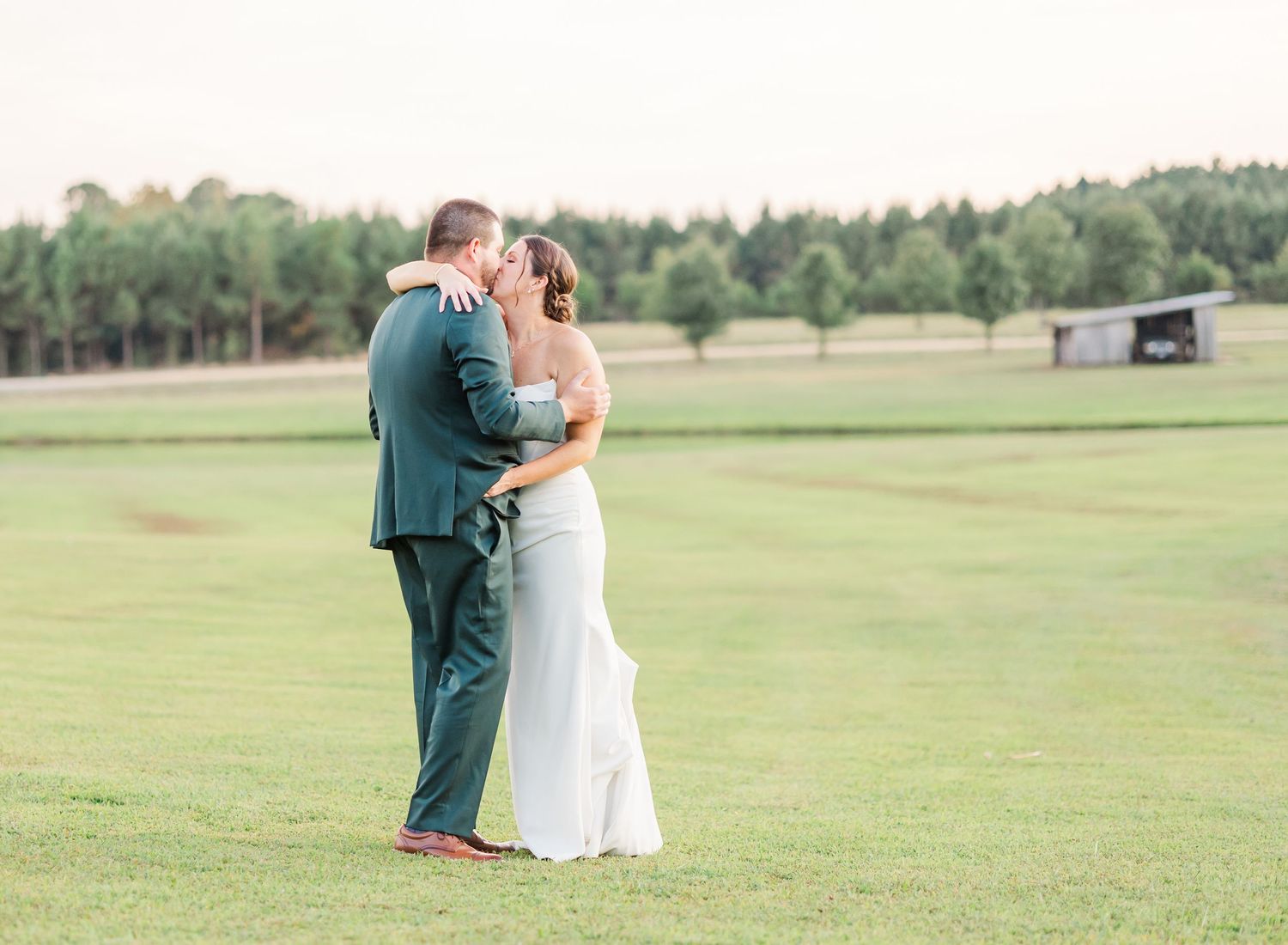 A couple shares an intimate moment on a lush green field with trees in the background on their wedding day.