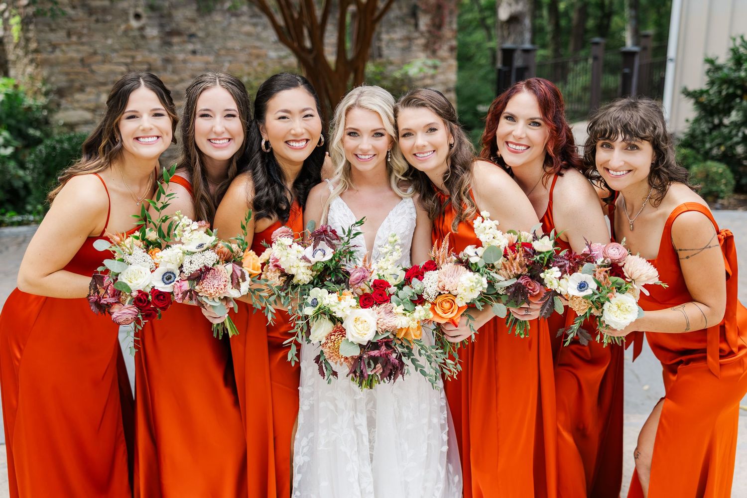 Bridesmaids in vibrant burnt orange dresses stand together holding colorful fall bouquets at an outdoor wedding.