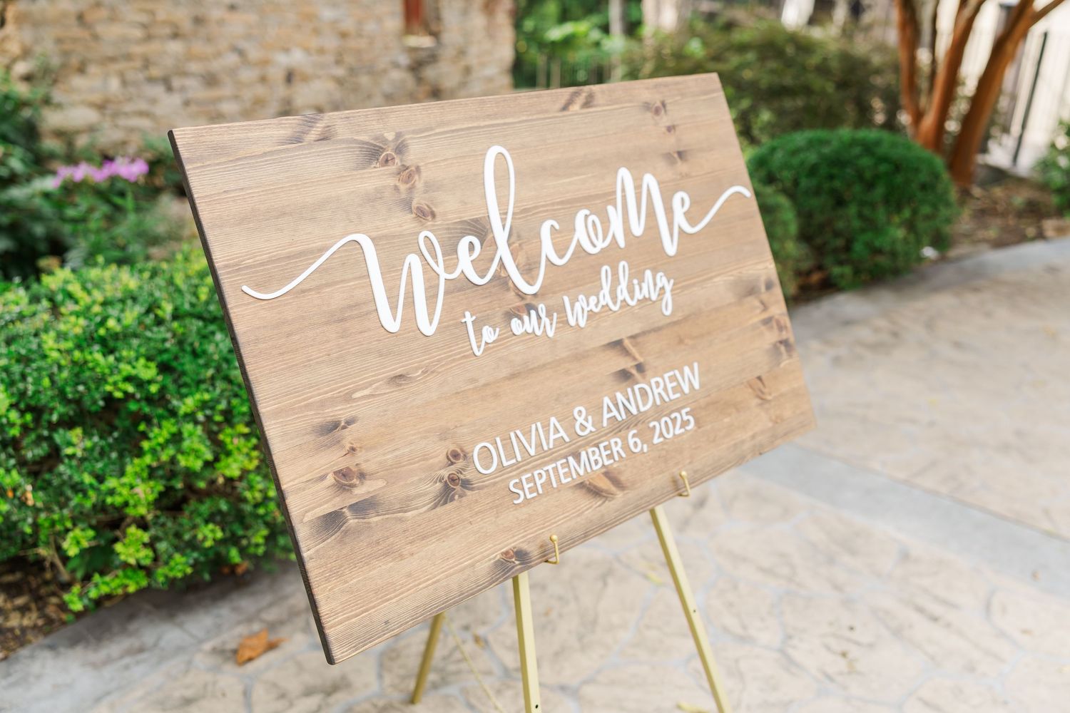 Rustic wooden wedding welcome sign with white calligraphy text stands on gold easel next to garden landscaping.