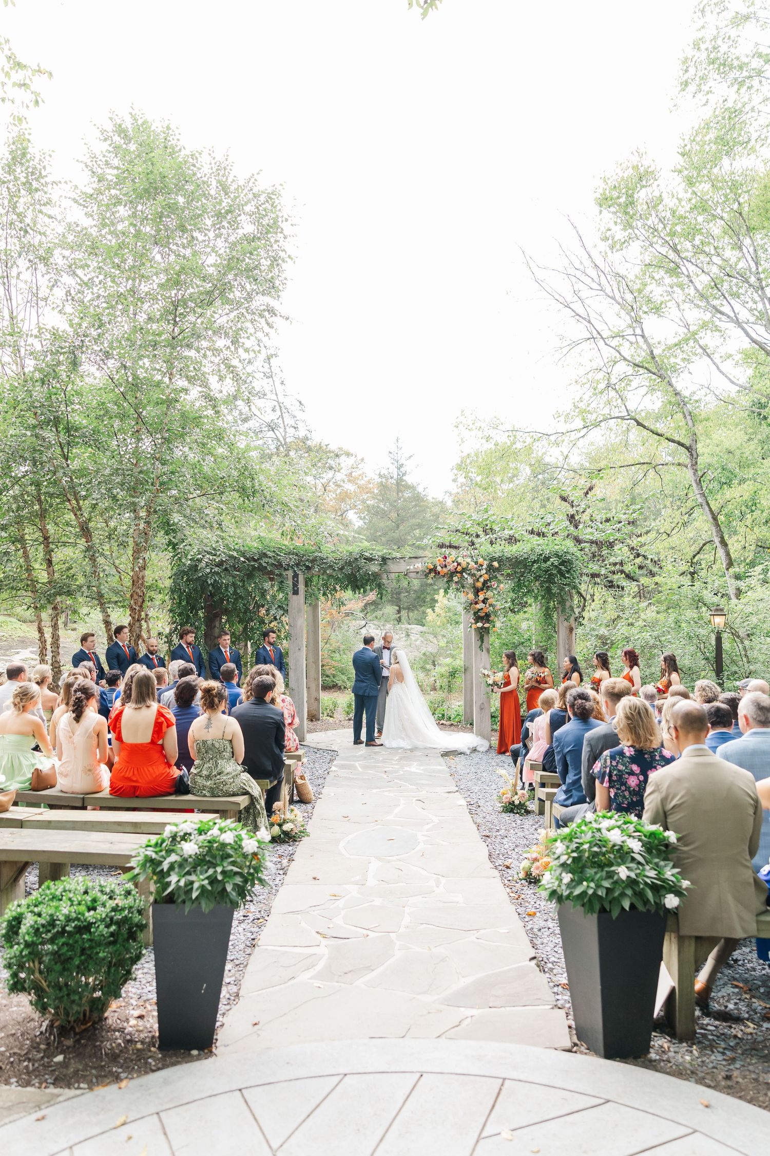 Wedding ceremony setup outdoors with guests seated in rows and large white floral arrangements in black planters.