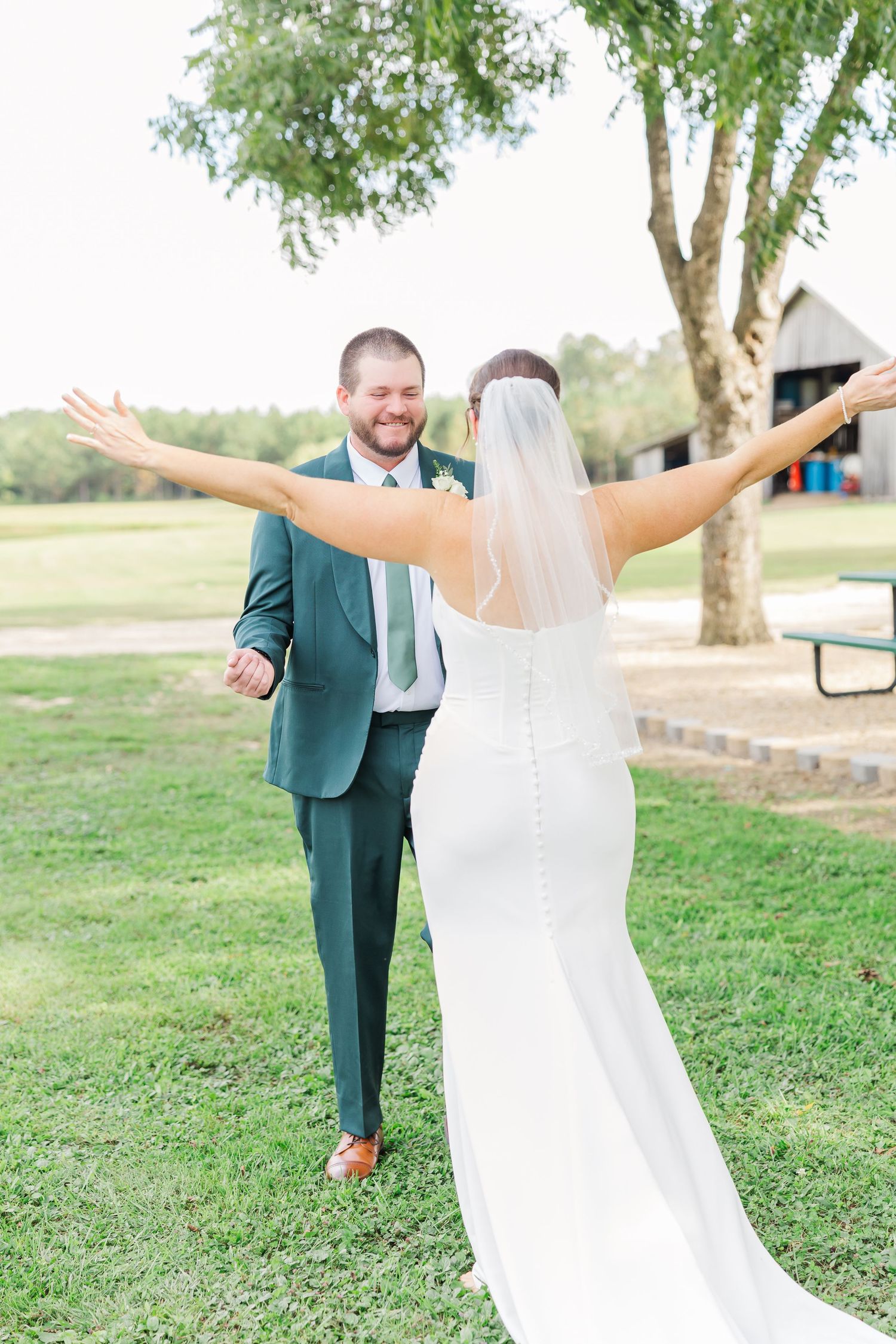 Bride in flowing white dress celebrating with arms outstretched on a sunny lawn.