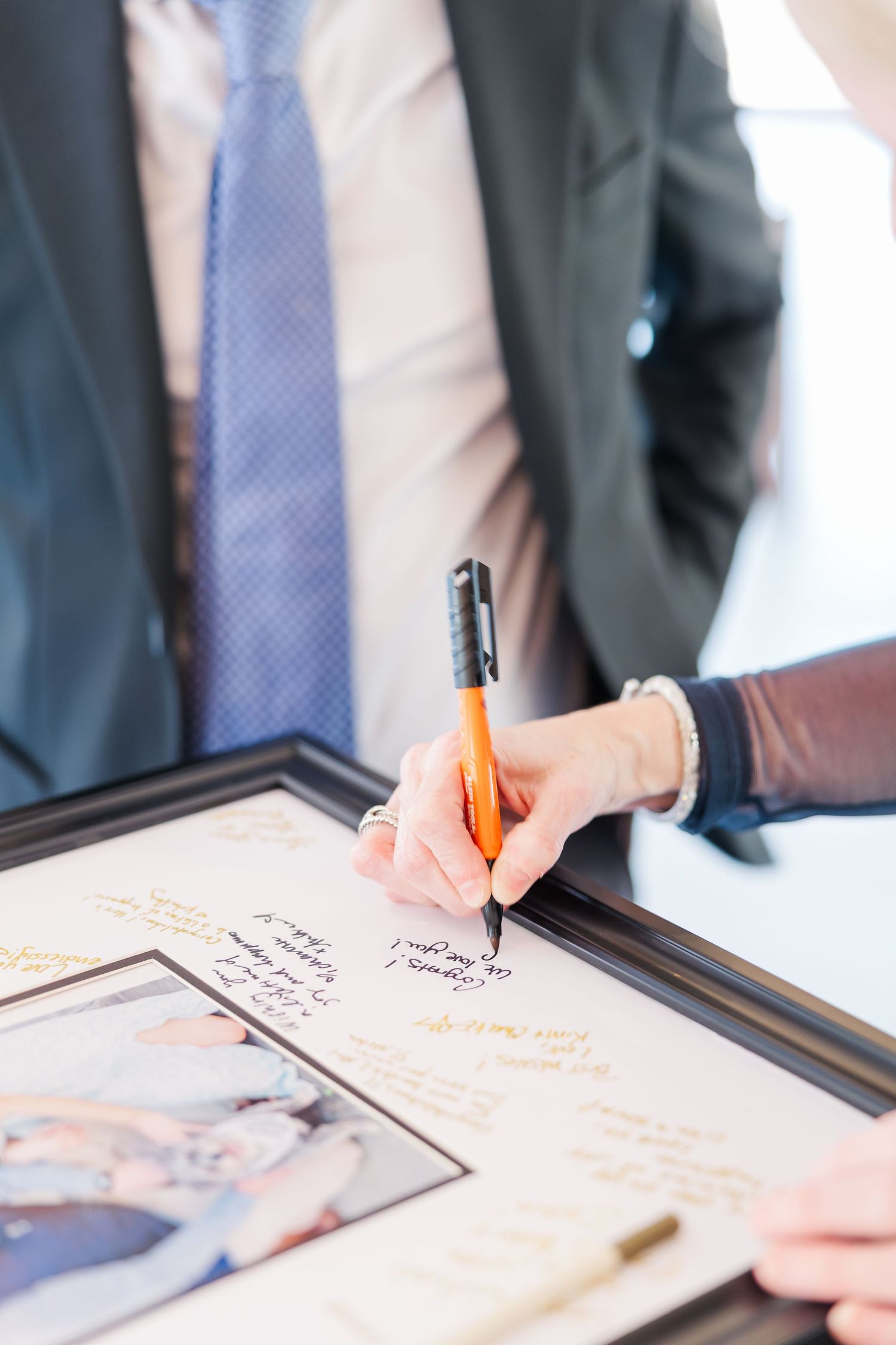 Hand signing a framed wedding guest book with an orange pen, man in suit with blue tie visible in background.