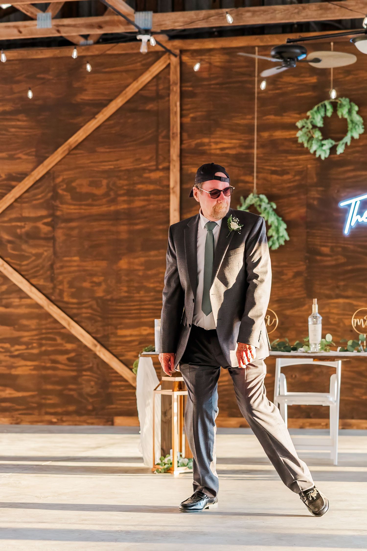 Groom in gray suit and sunglasses walking confidently at rustic barn wedding venue with string lights.