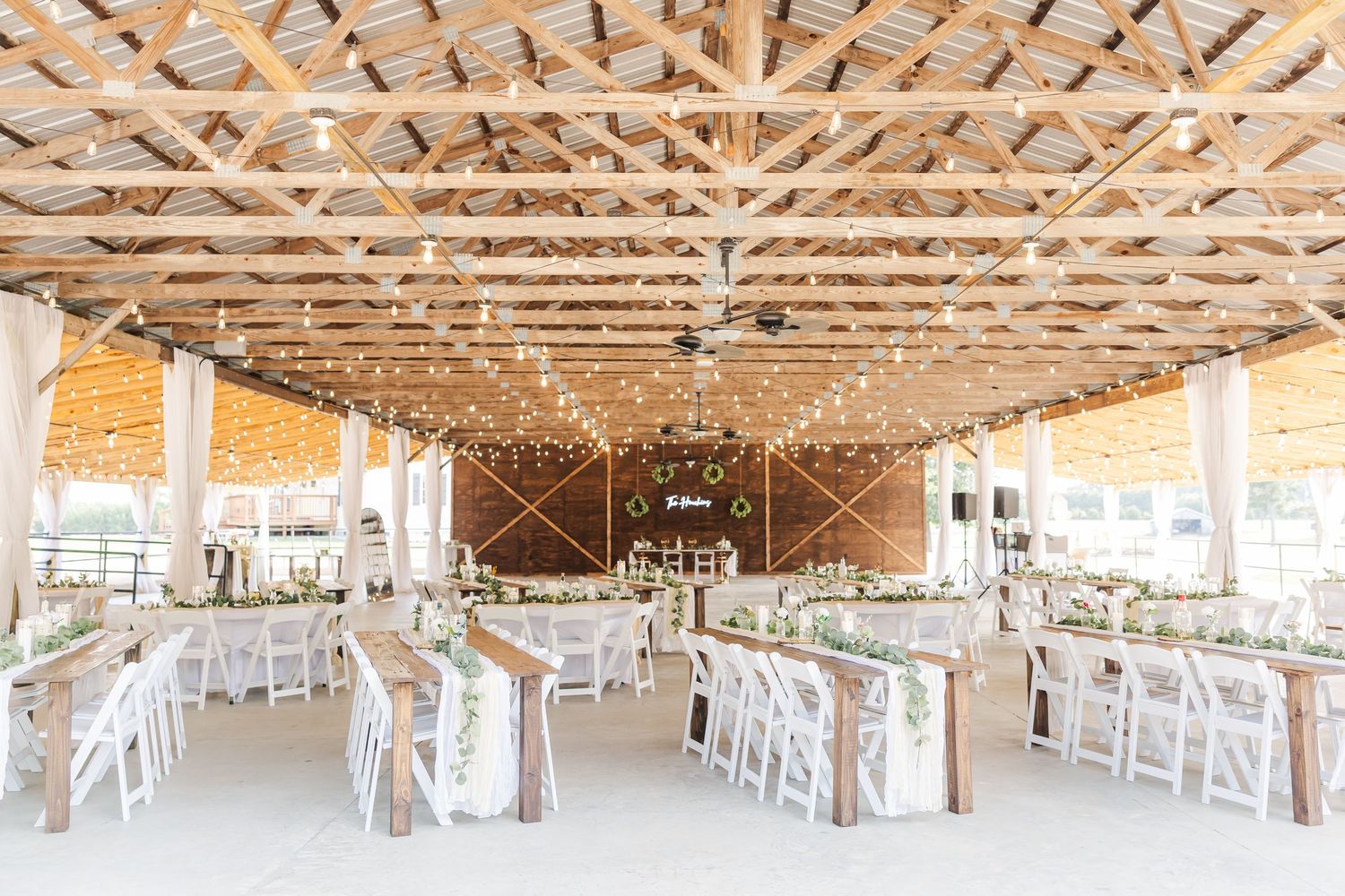 Rustic barn wedding reception with string lights, wooden beamed ceiling, long farm tables, white chairs, and greenery centerpieces.