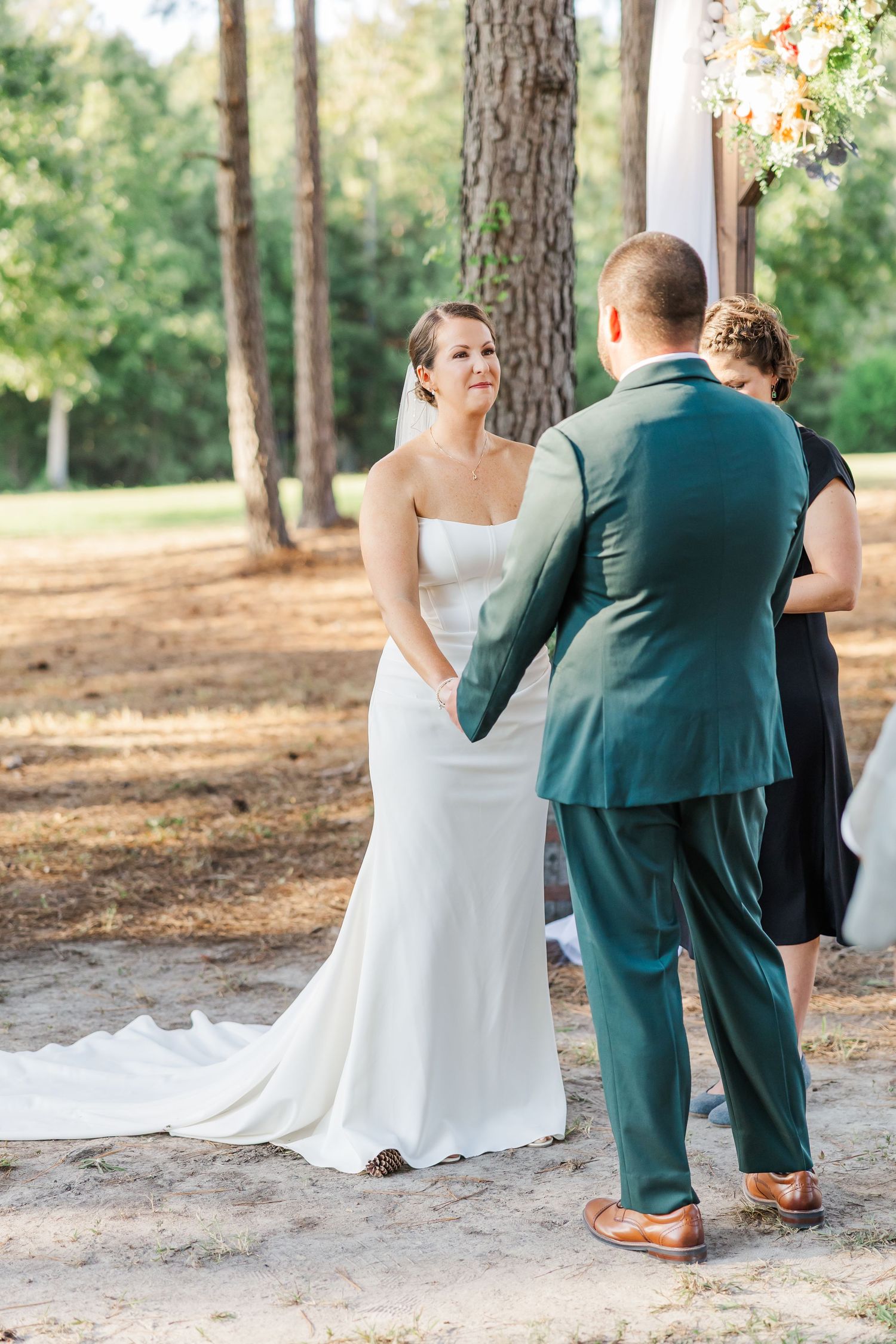 Bride in white gown holds grooms hands during outdoor wedding ceremony in pine forest, officiant stands behind couple.