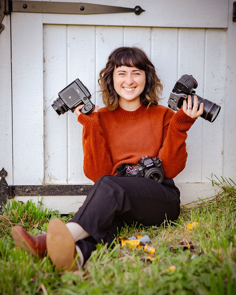 Photographer wearing orange sweater poses with cameras against white barn door.