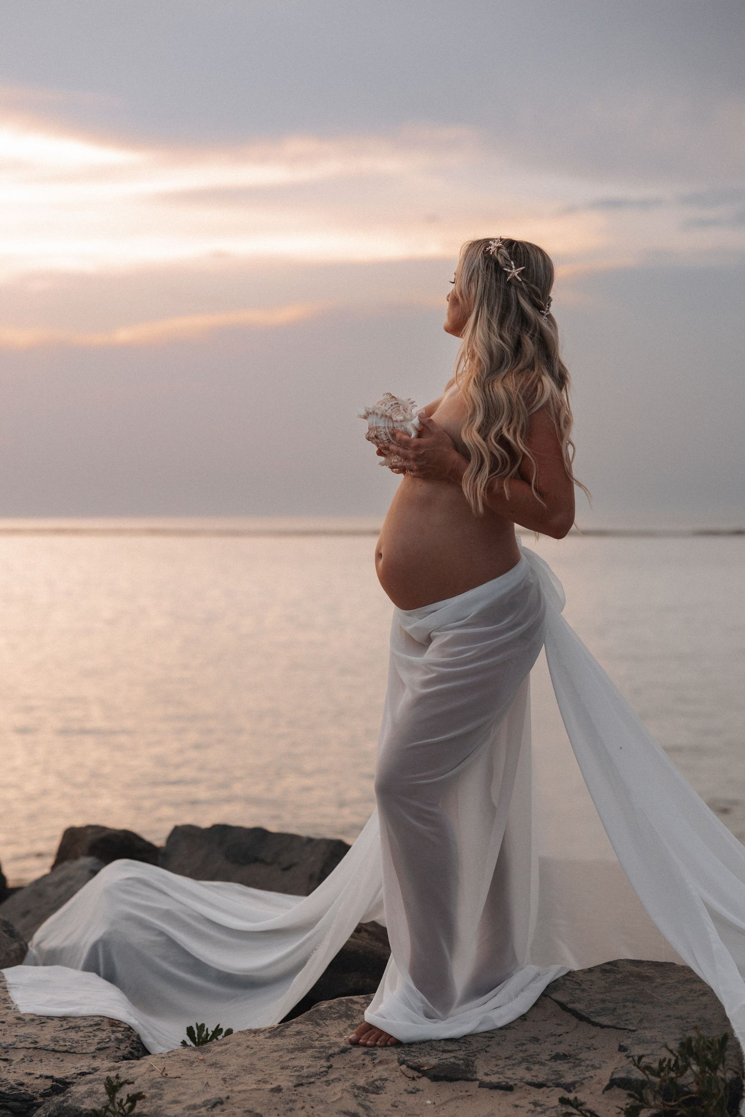 Artistic maternity photos at sunset with flowing white fabric on a beach.