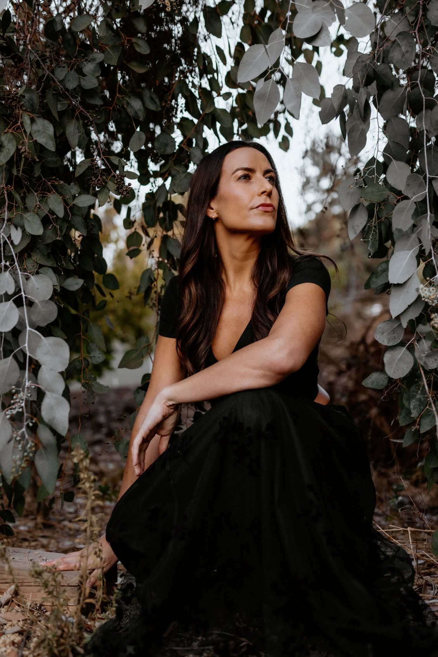 A woman in a black dress poses against a backdrop of hanging leaves in moody outdoor lighting.