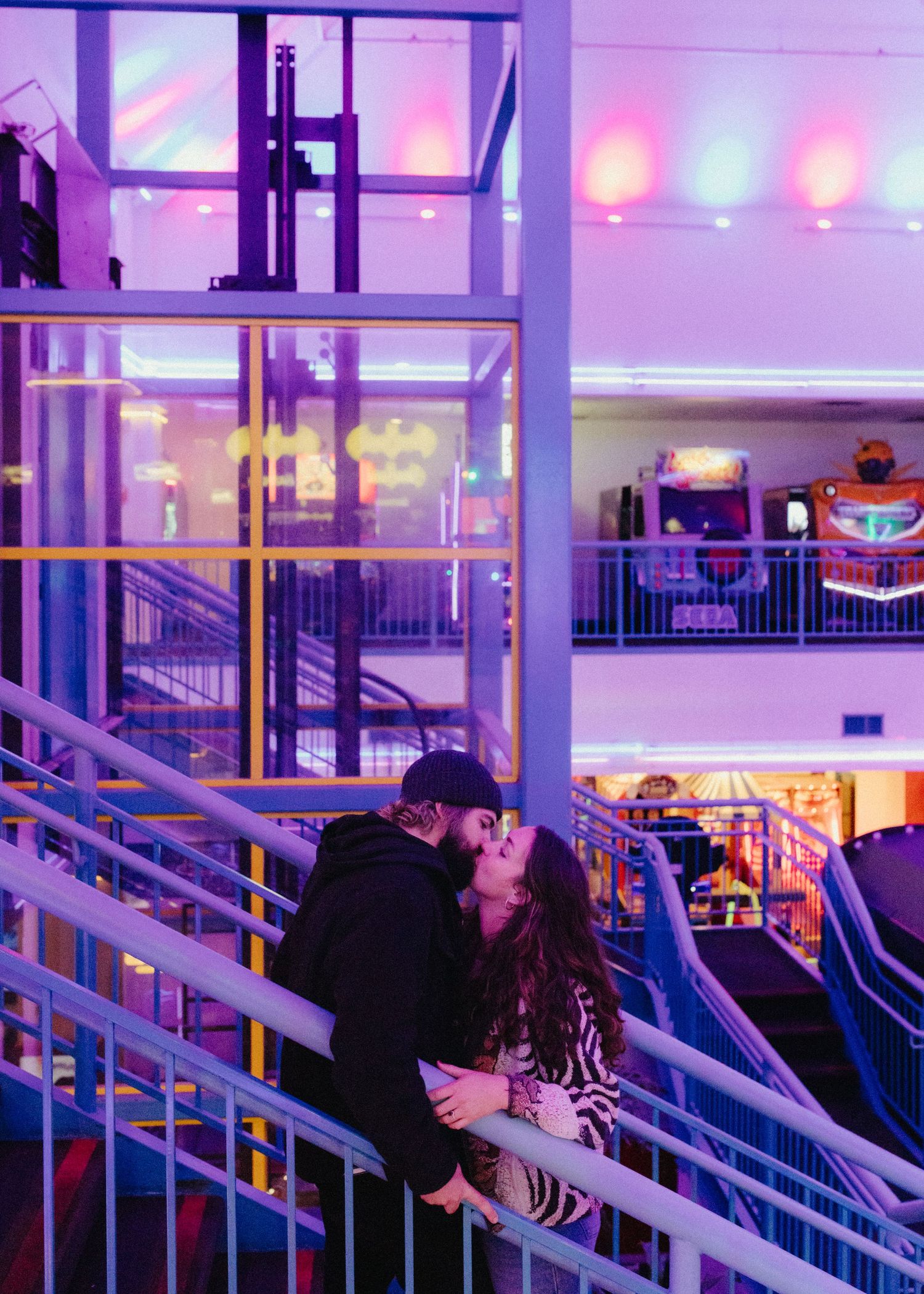 Colorful LED lights illuminate a modern mall interior with glass elevators and escalators creating a vibrant neon atmosphere.