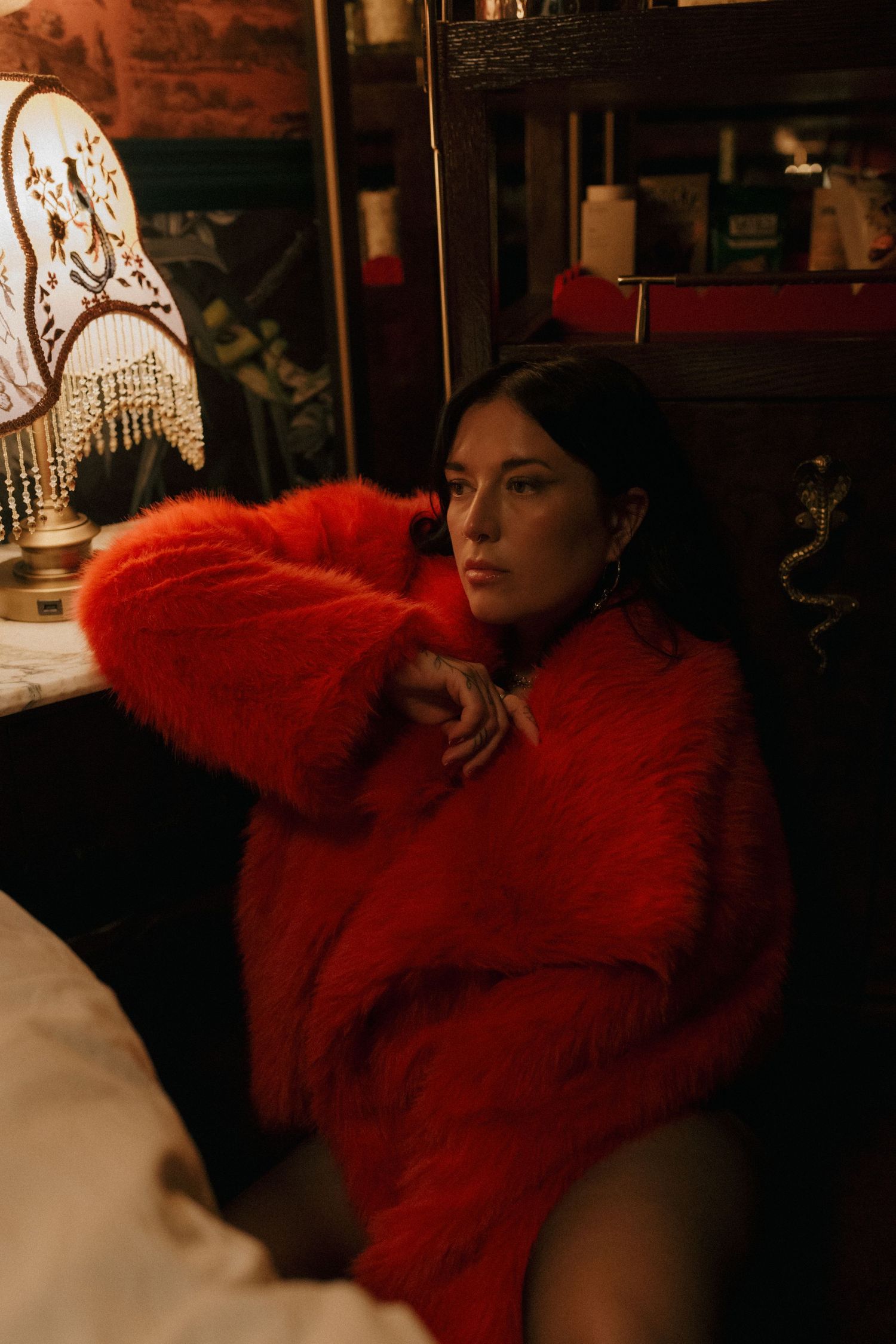 Moody portrait in dark room featuring dramatic red furry coat against vintage-style decor and lampshade.