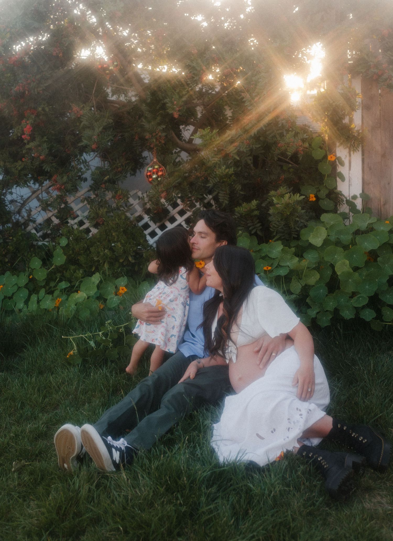 A dreamy garden scene with people sitting on grass surrounded by lush greenery and sunlit trees during golden hour.