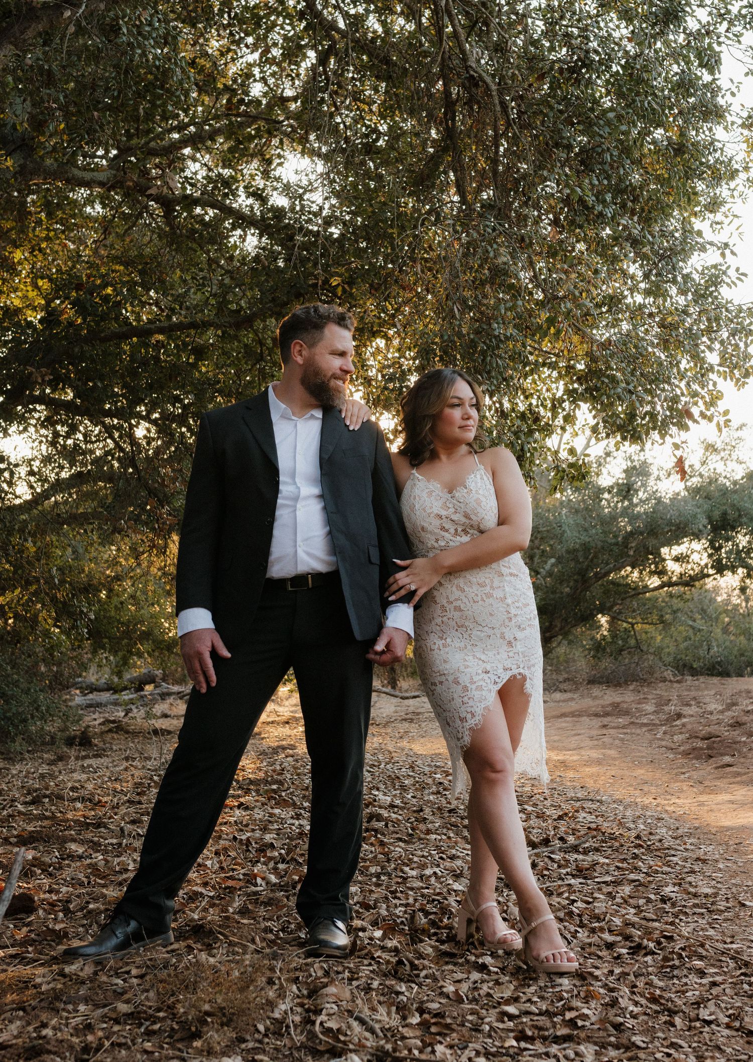 A romantic outdoor photoshoot featuring a couple in formal attire surrounded by trees at sunset.