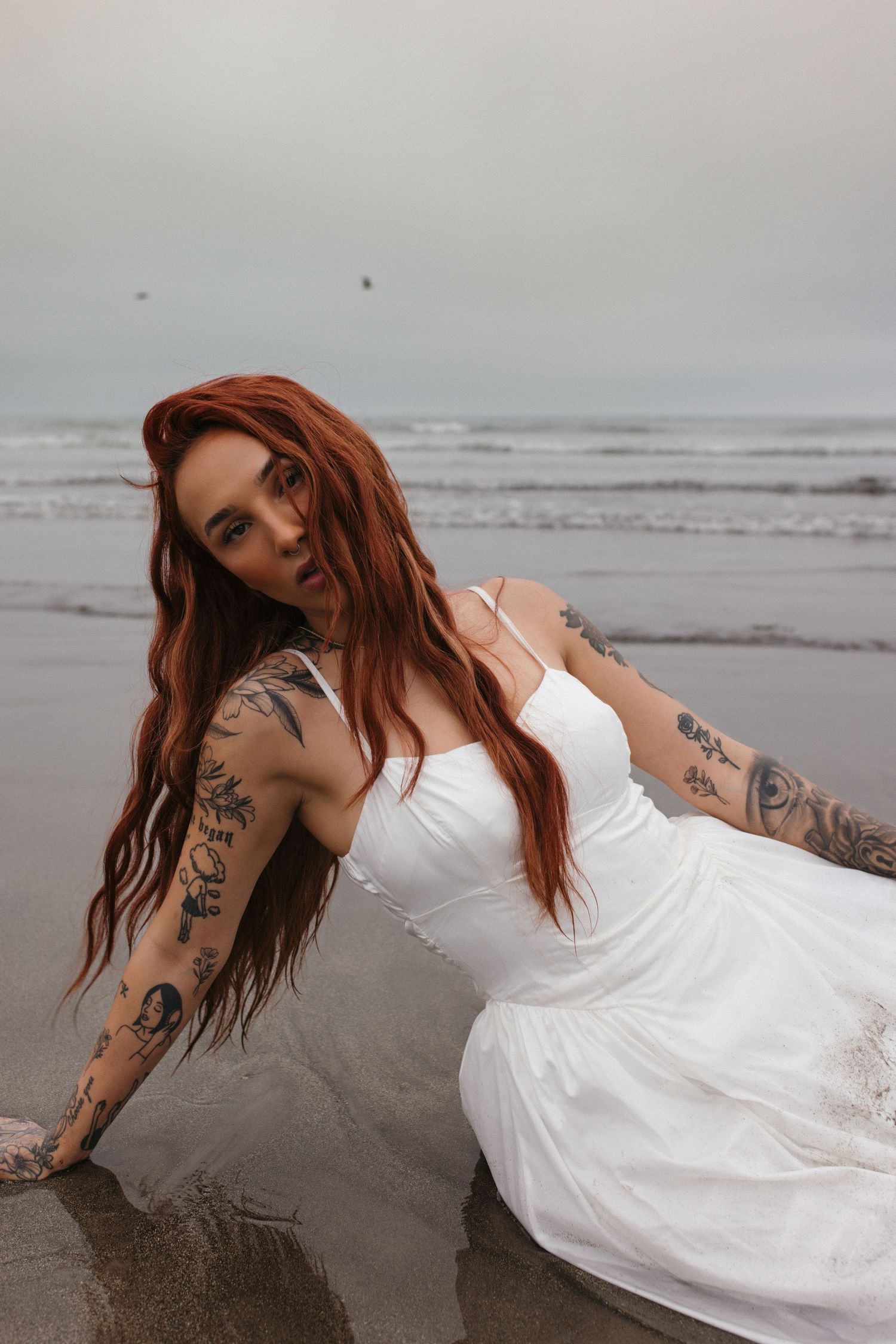 Woman in white wedding dress with tattoos poses on misty beach.