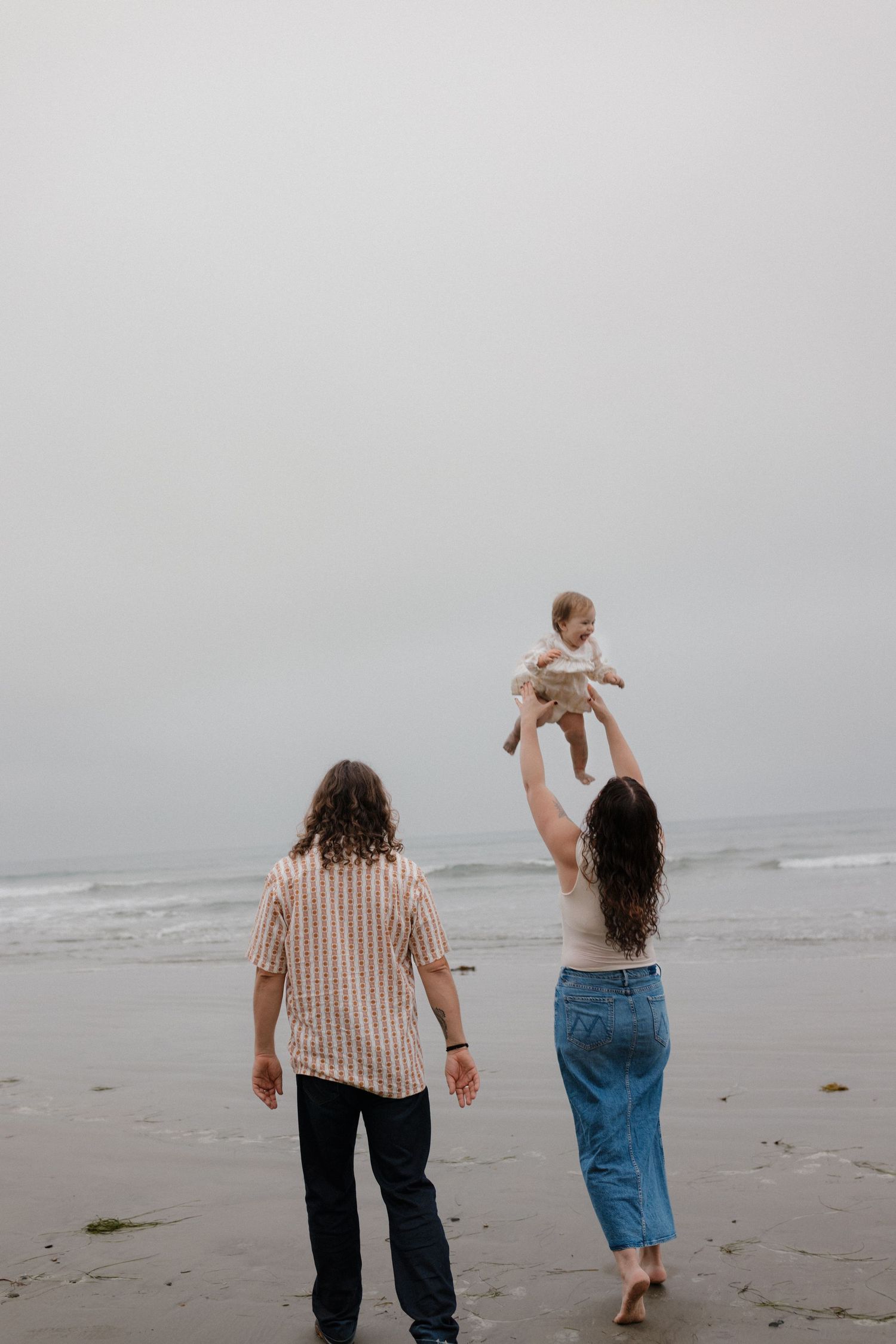 Family enjoying a day at a foggy beach with child being lifted into the air.