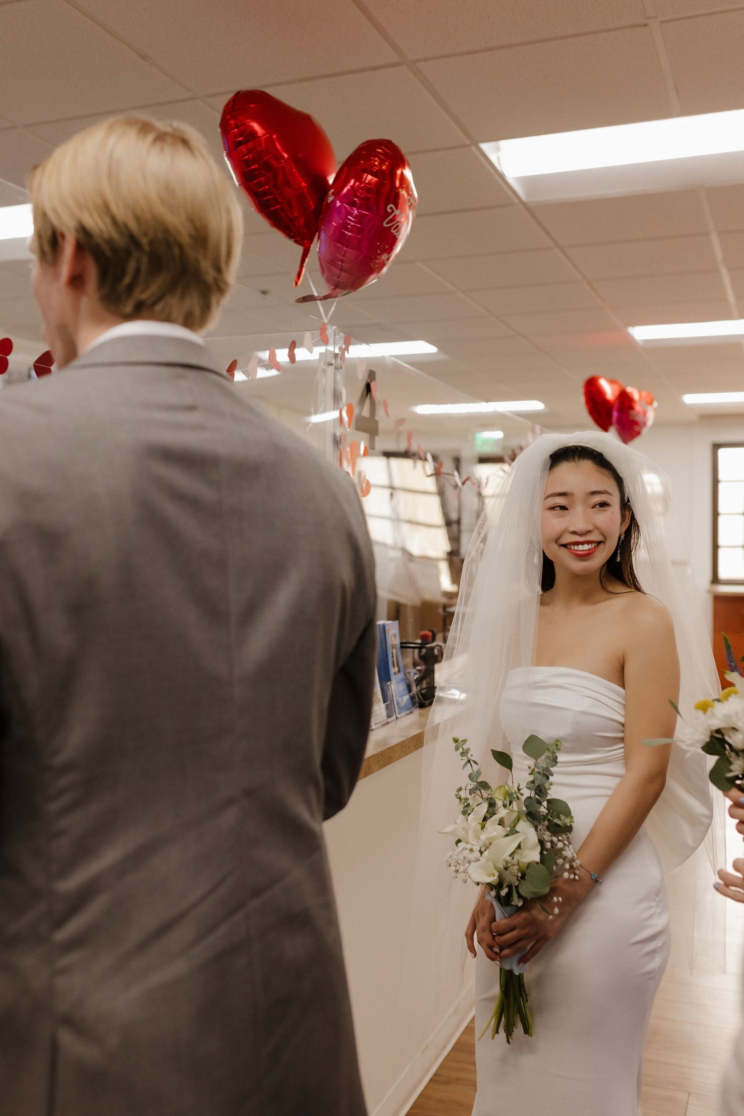 A wedding couple holds heart-shaped balloons in an indoor hallway setting with simple elegant decorations.