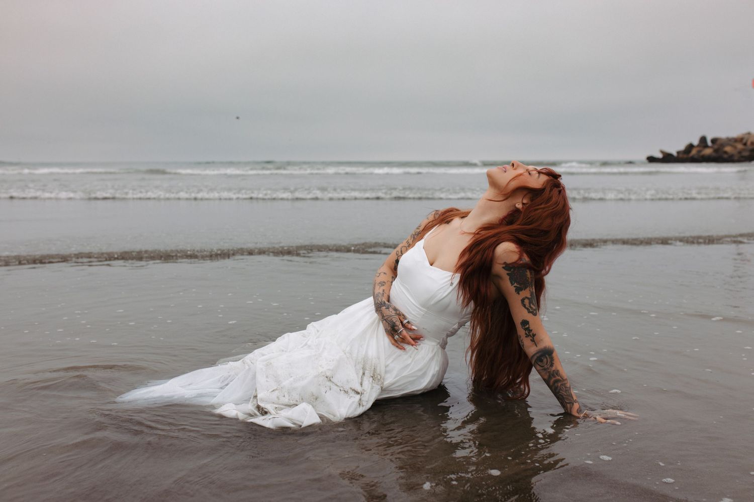 Woman in white wedding dress poses dramatically on wet sand at beach.