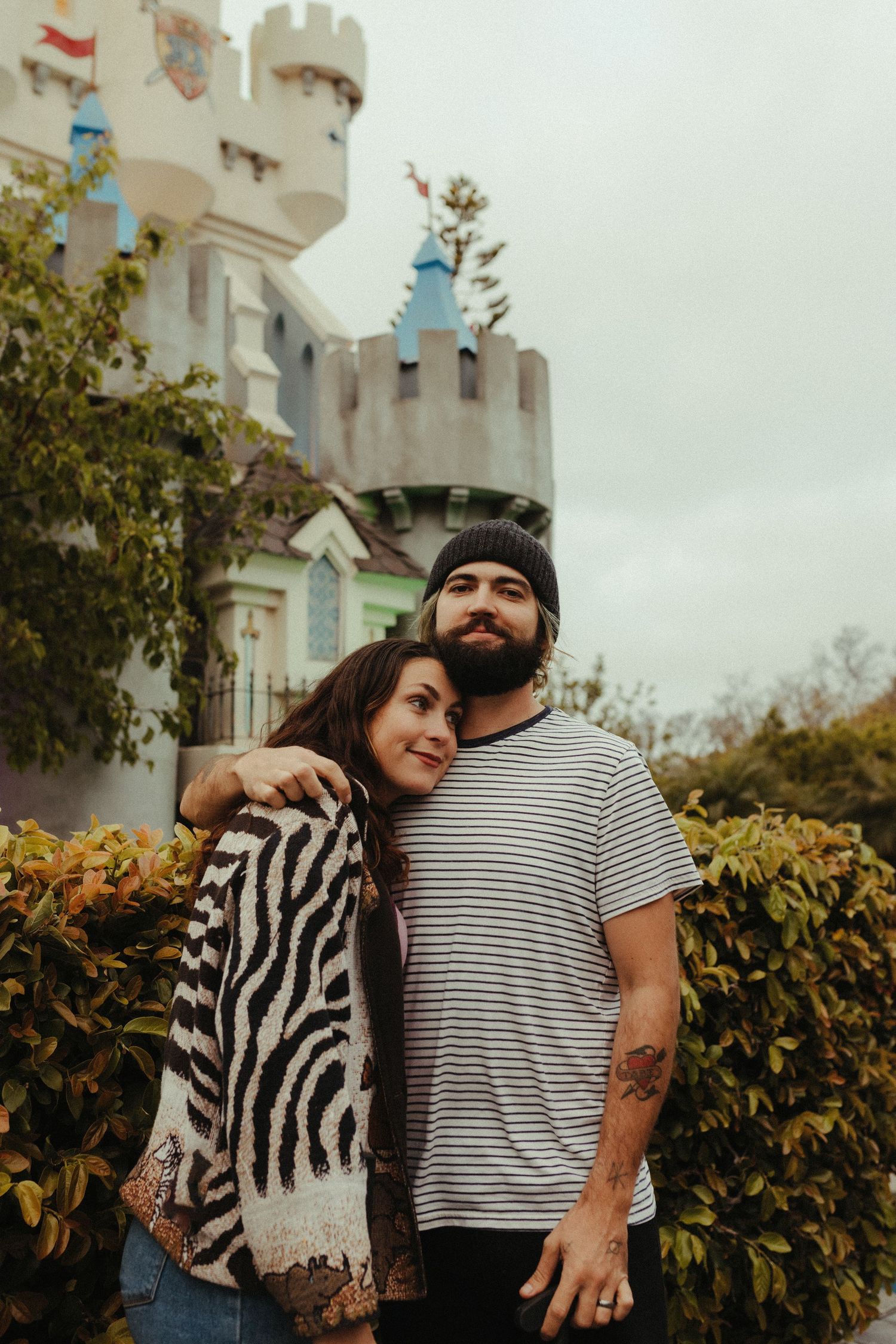 A couple stands together in front of a castle-like building with turrets on an overcast autumn day.