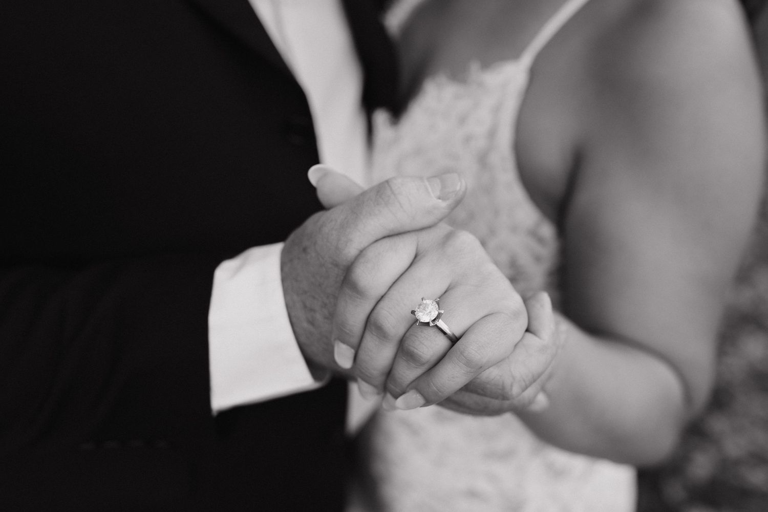 Black and white close-up of clasped hands showing an engagement ring during a wedding ceremony.