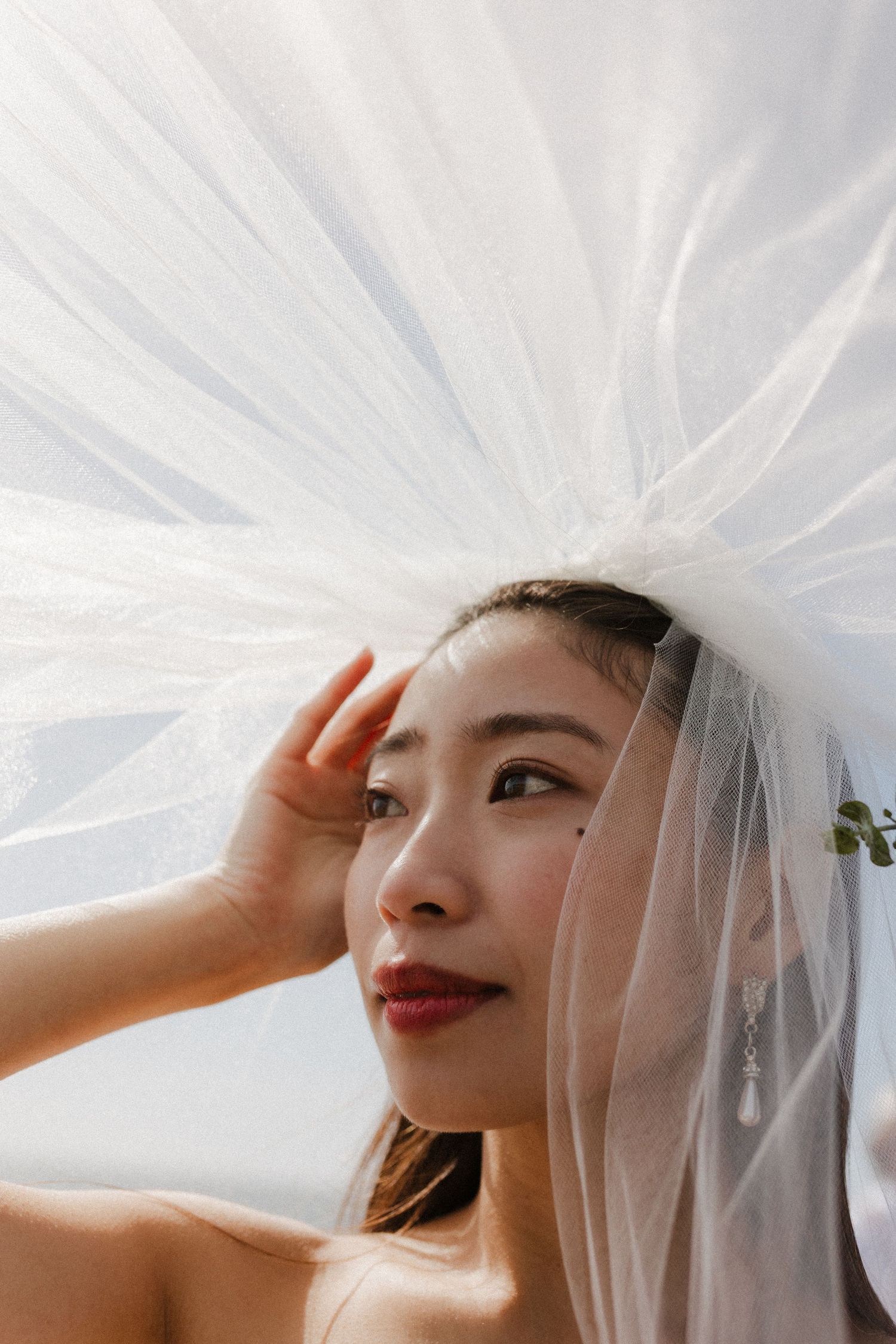 A bride in a white veil poses in soft sunlight wearing red lipstick against a light background.