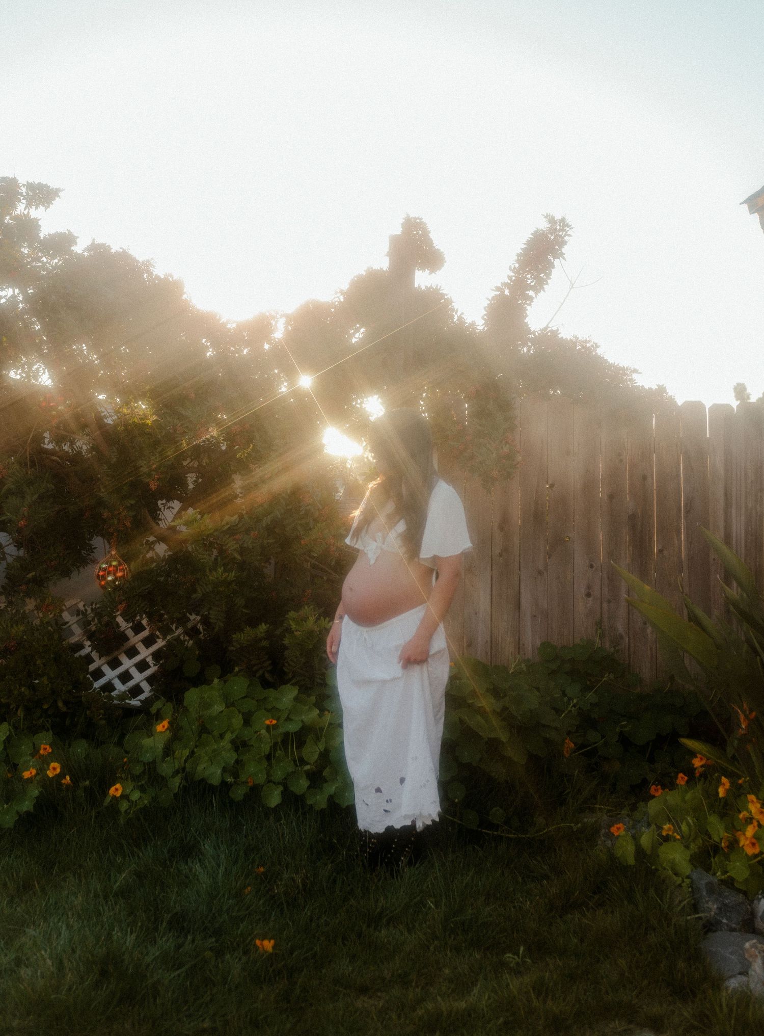 Figure in a white dress stands in a garden at sunset with sunbeams streaming through trees and orange flowers blooming.