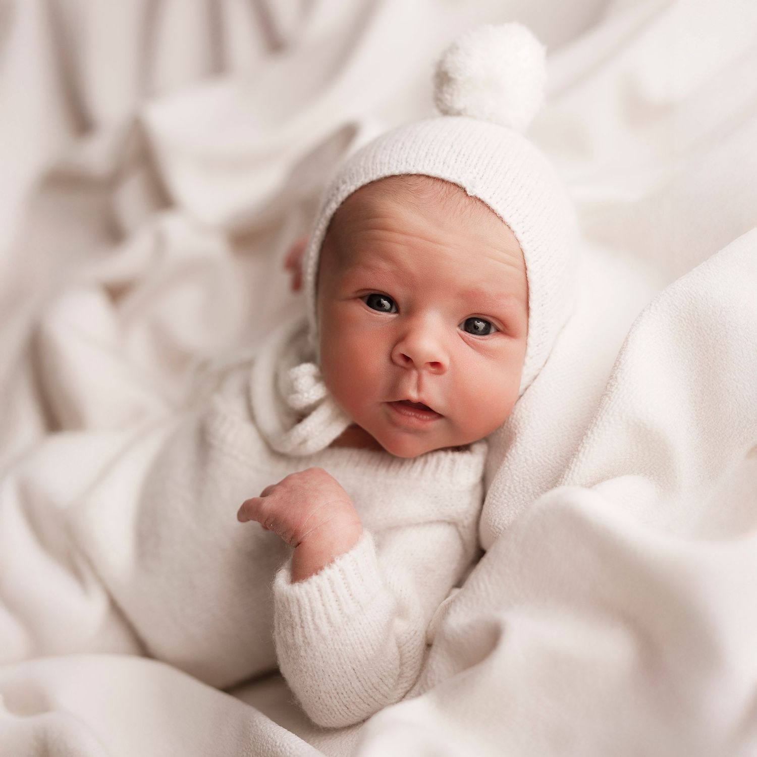 Sweet newborn baby wearing white knit clothing rests peacefully on soft white bedding in a series of gentle poses.