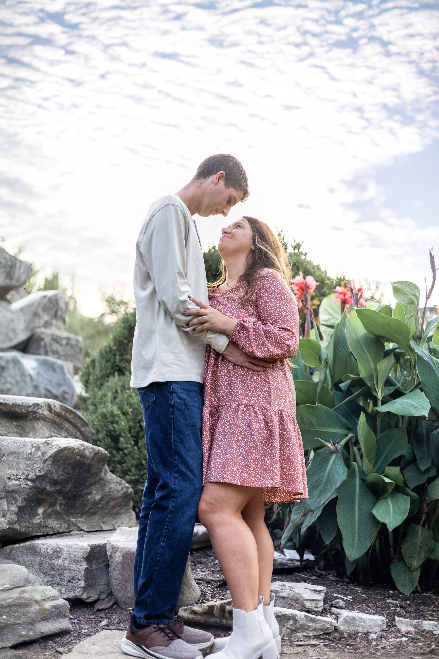 Romantic couple embracing on rocky outcrop with lush plants and cloudy sky.