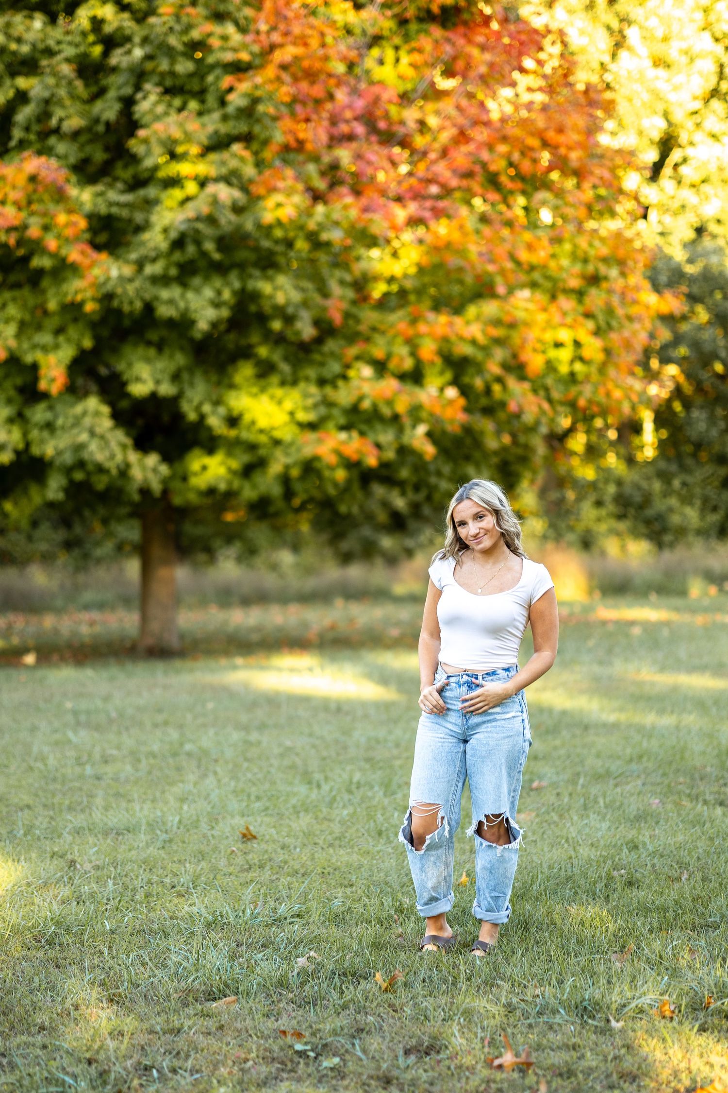 Woman in casual attire sitting on grass during autumn with colorful fall foliage in background.