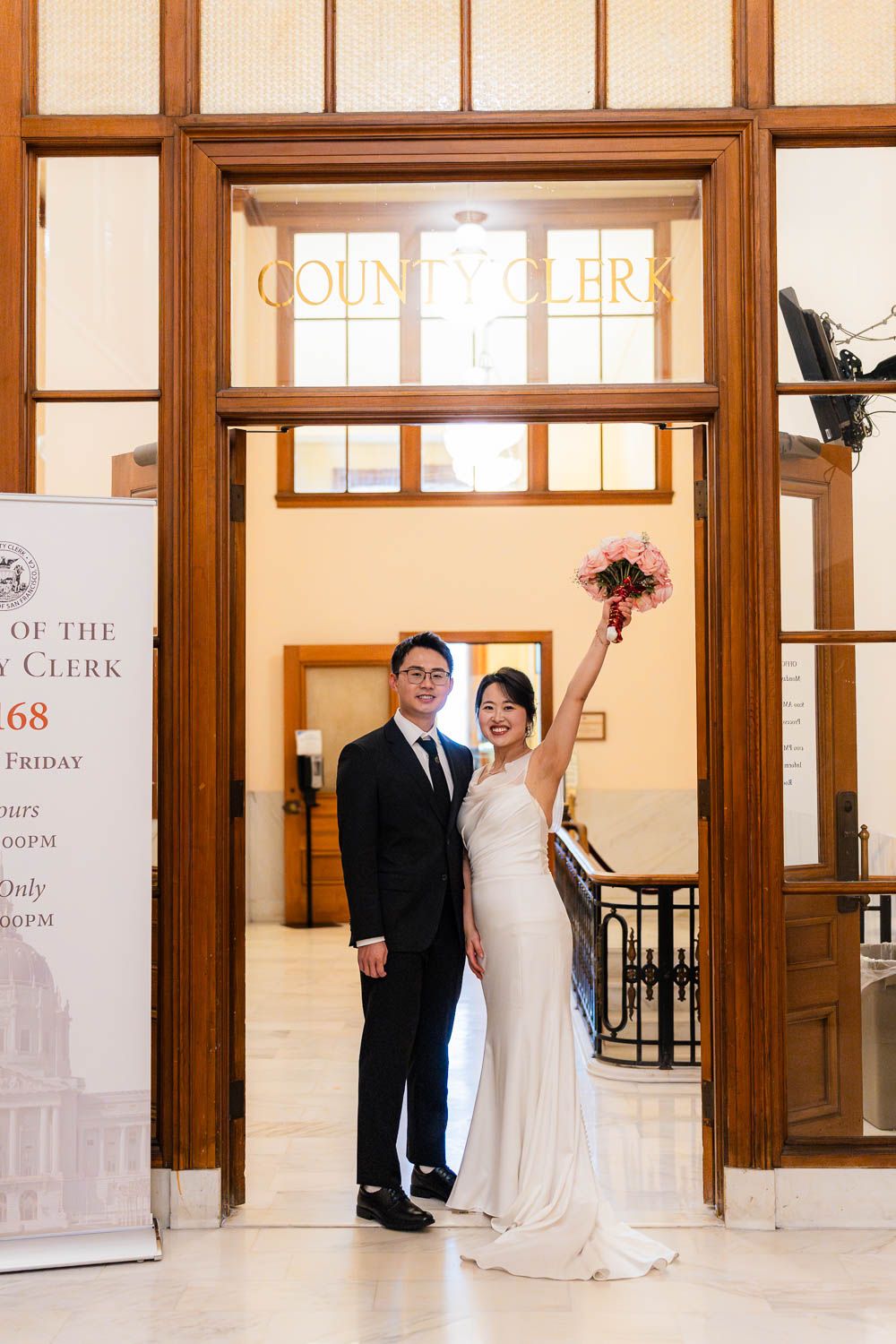 A couple in wedding attire stands in front of elegant wooden doors at County Clerk's office in San Francisco City Hall.