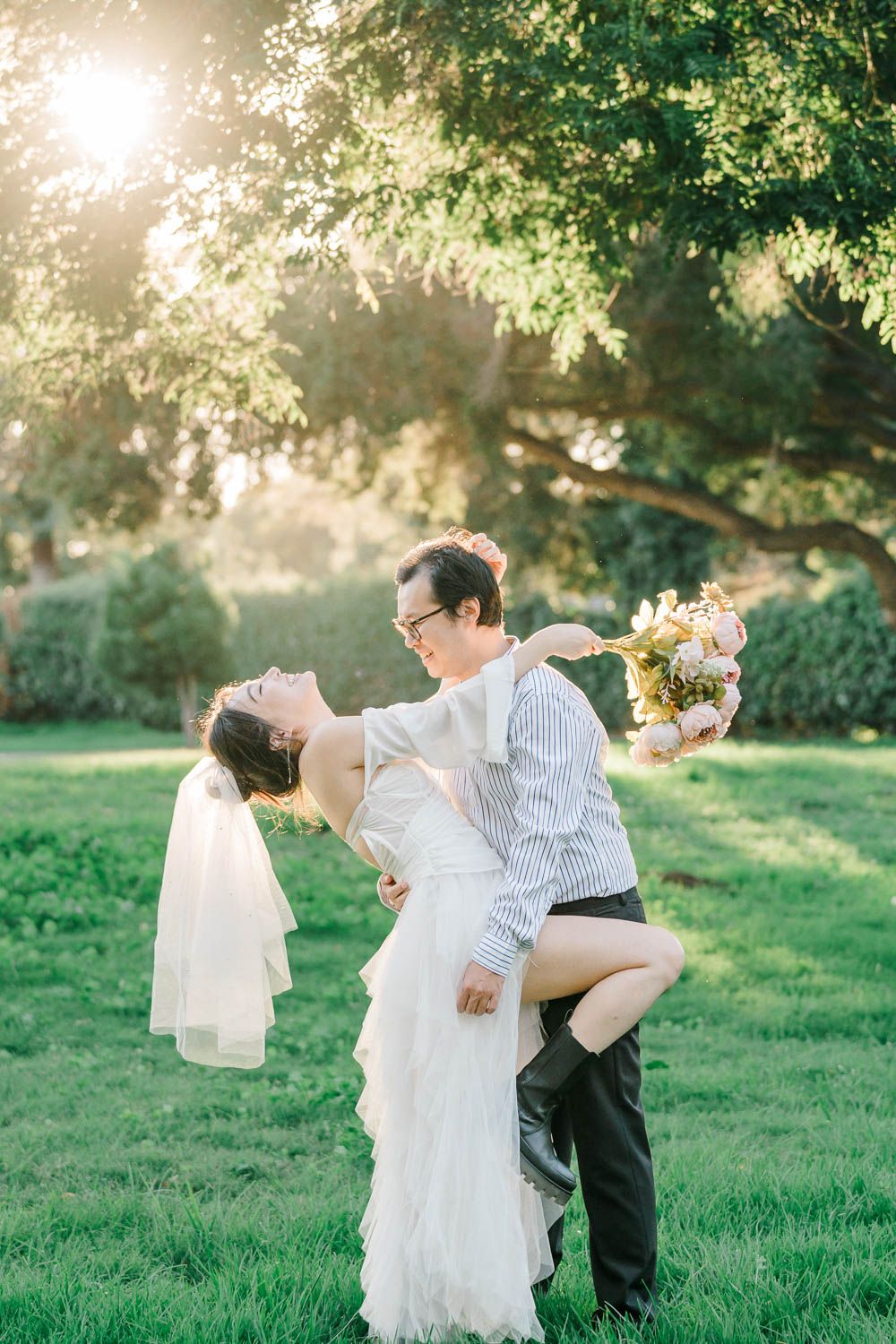 A couple dances together on grass in golden sunlight while wearing wedding attire.