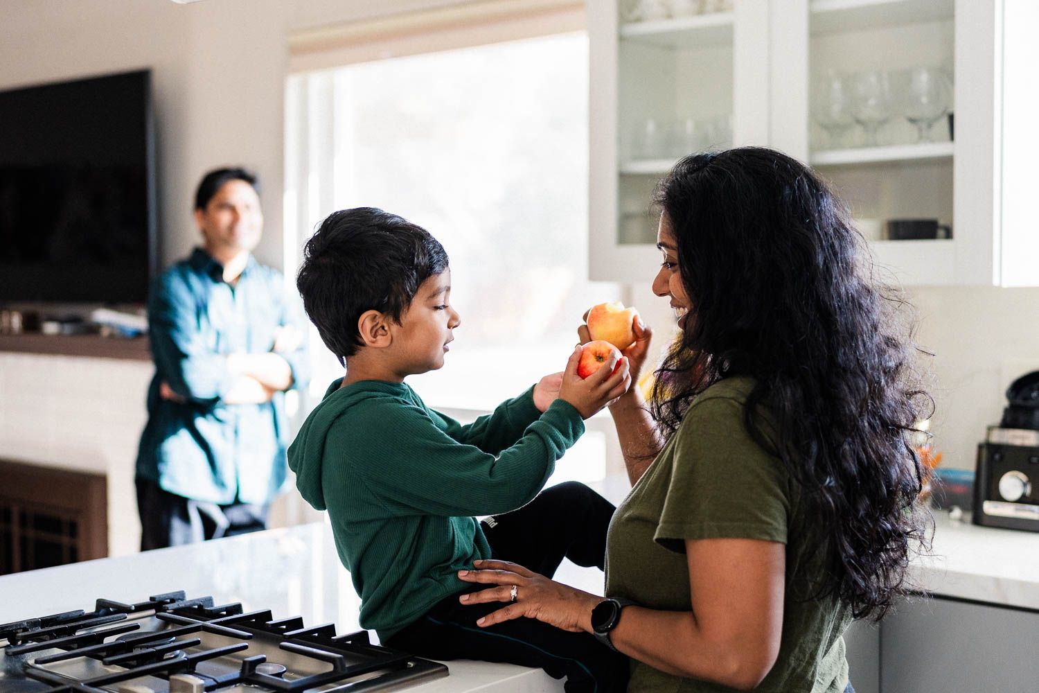 A young child shares an apple with mom in a modern kitchen while dad stands in the background looking at them.