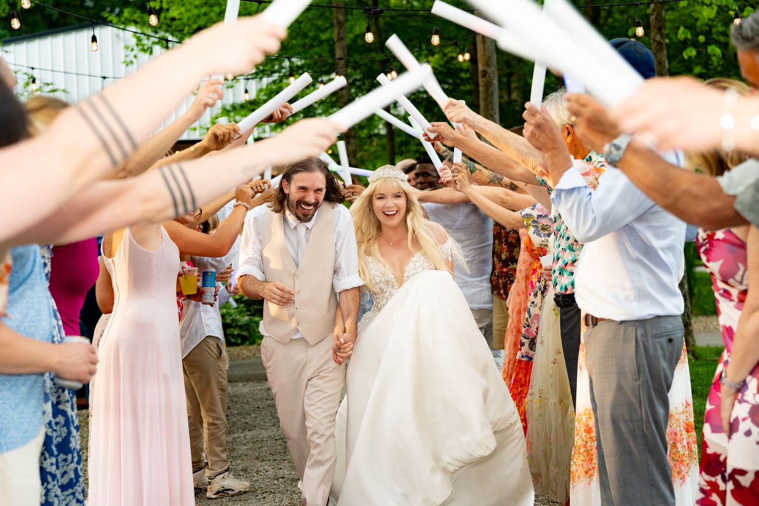 Wedding guests form a tunnel with raised arms as couple walks through during outdoor garden celebration.