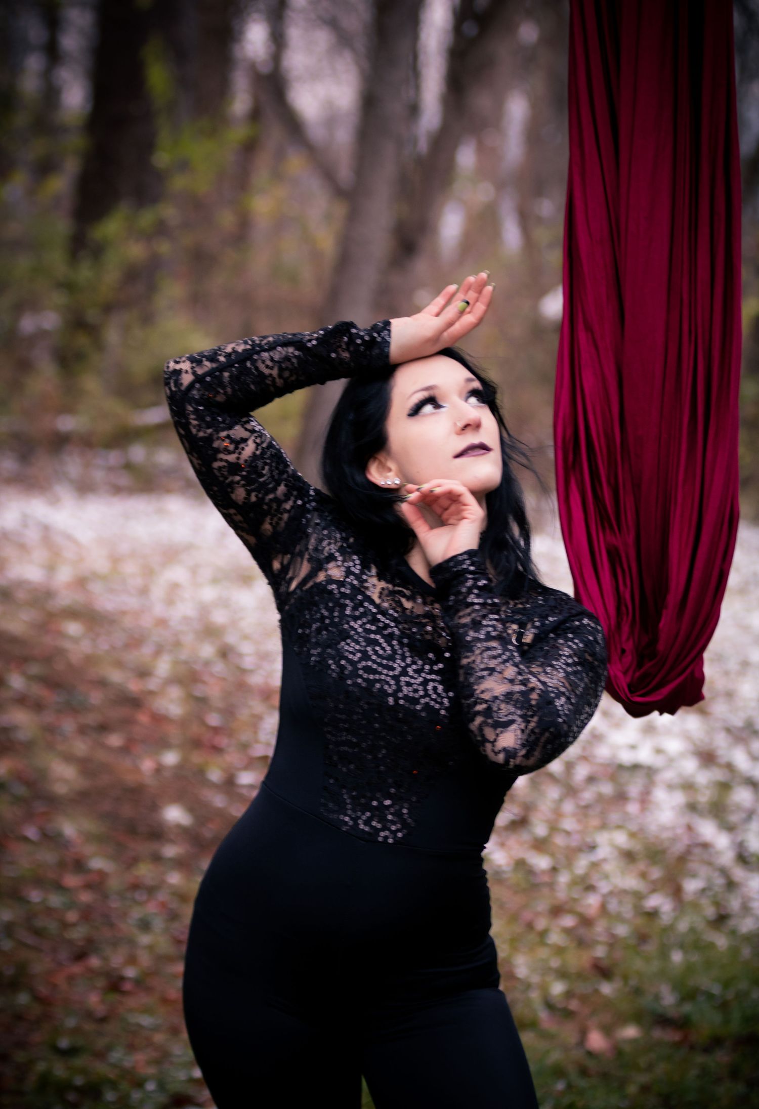 A dancer in a black lace dress performs an elegant pose against a burgundy fabric backdrop in a forest setting.