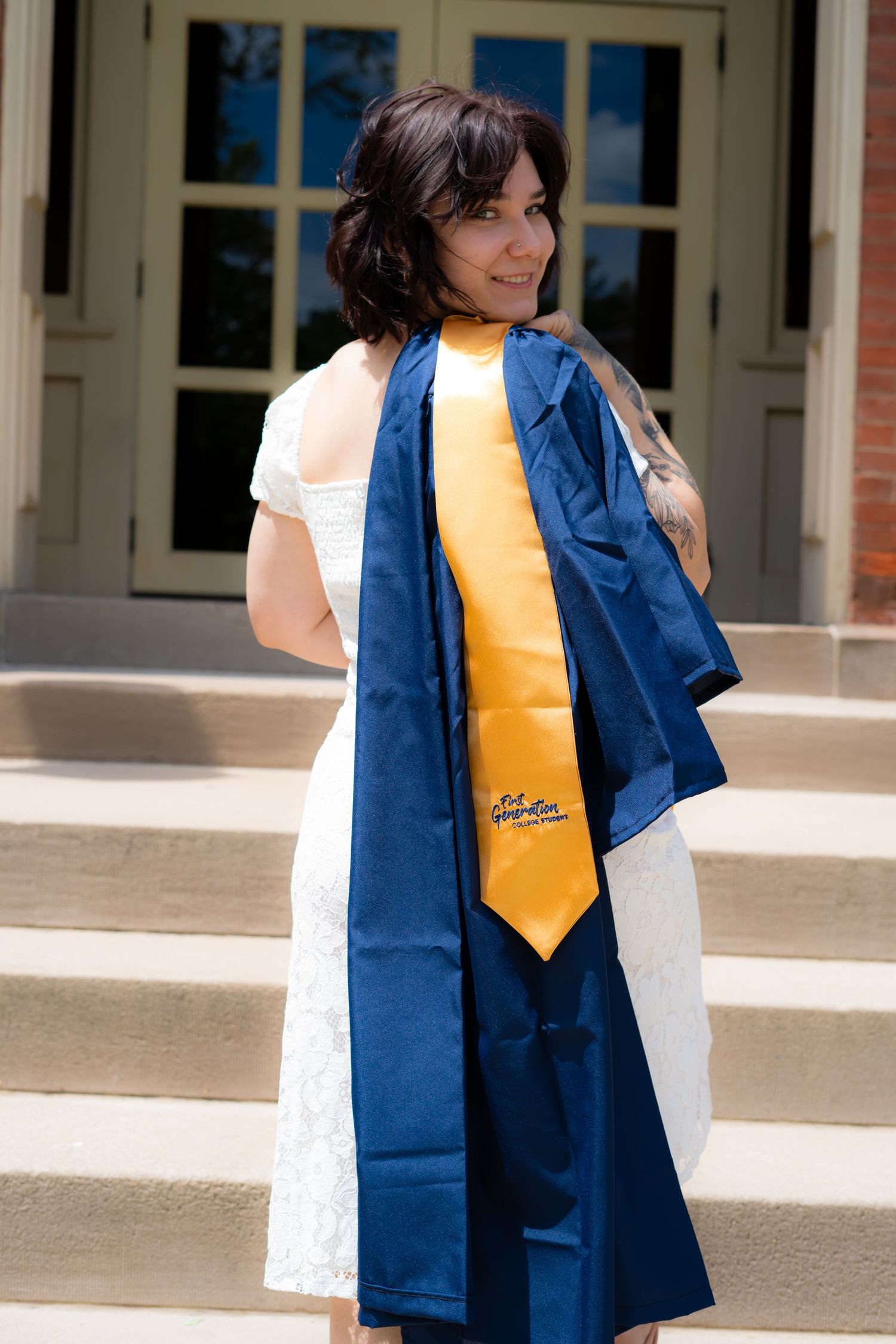 Graduate wearing blue cap and gown with gold tie poses on campus steps.