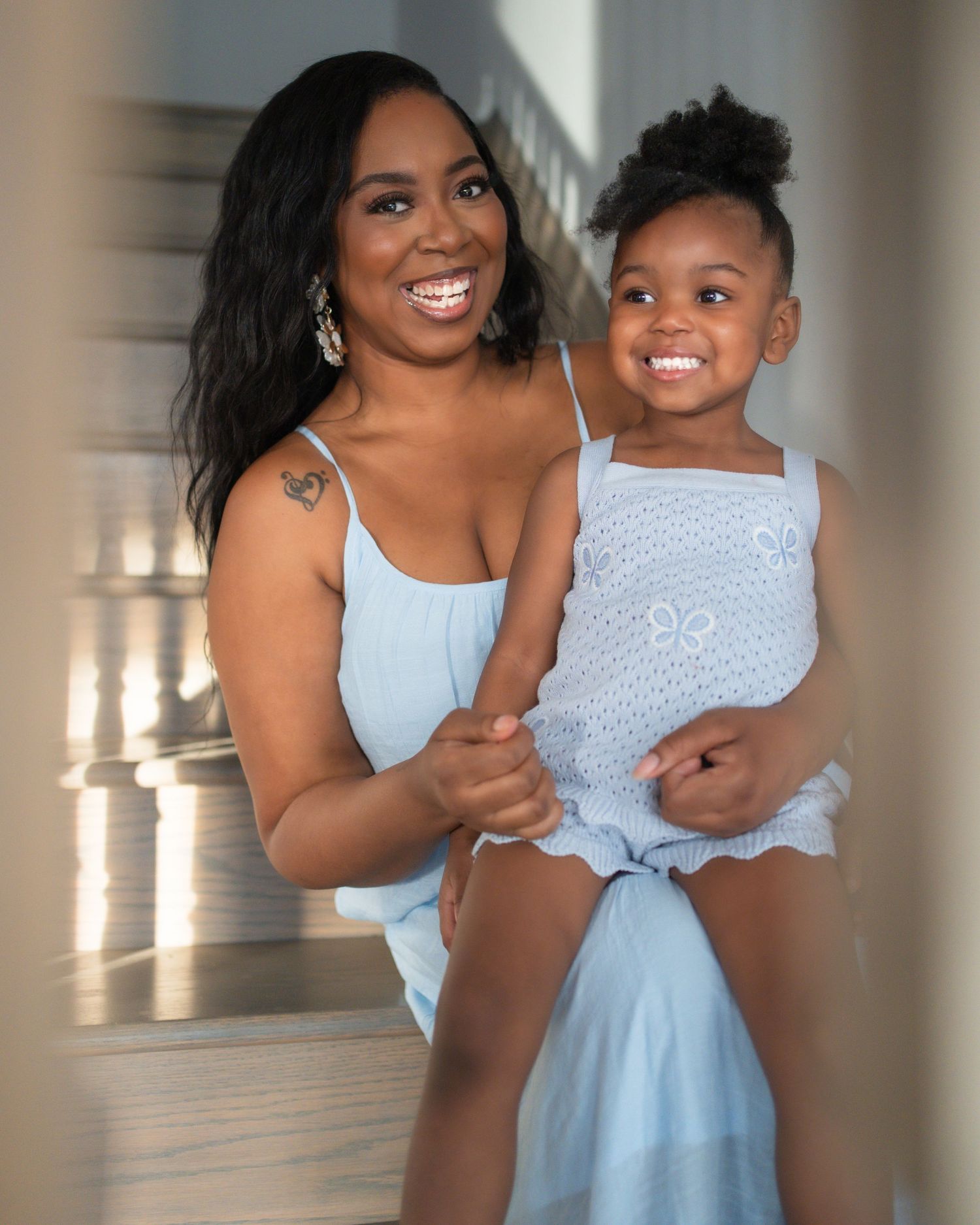 A smiling woman in a light blue dress holds a happy toddler girl in a matching blue outfit on stairs.
