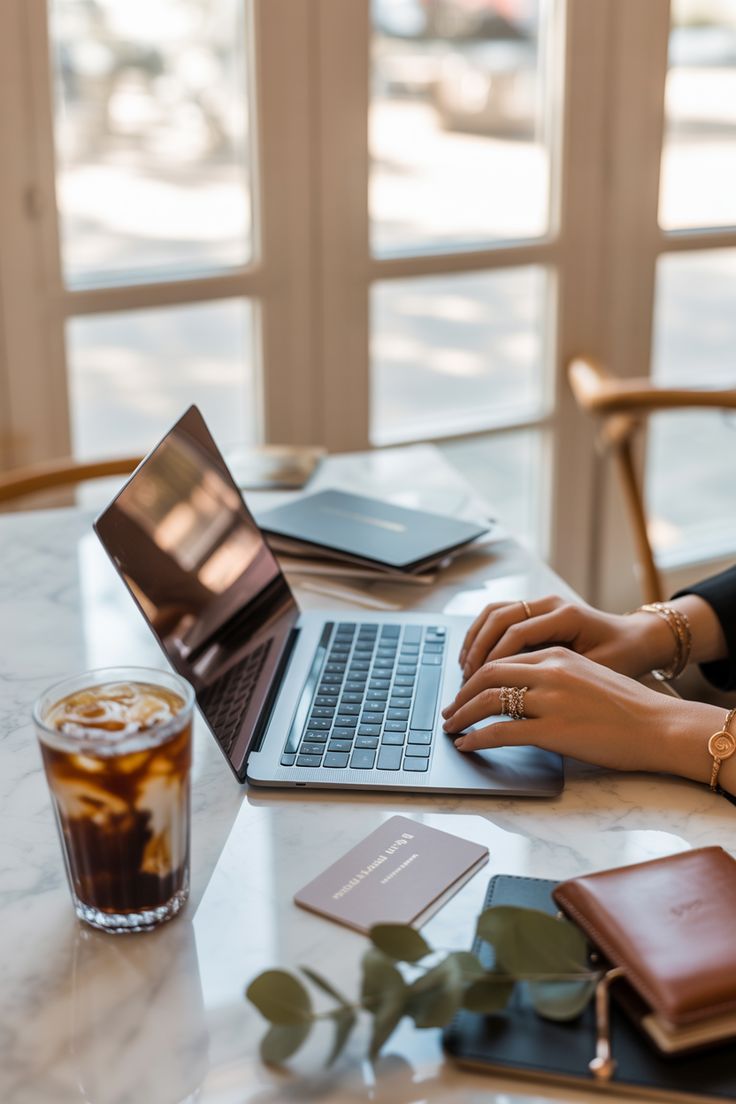 Hands typing on laptop with coffee and wallet on wooden desk by window.