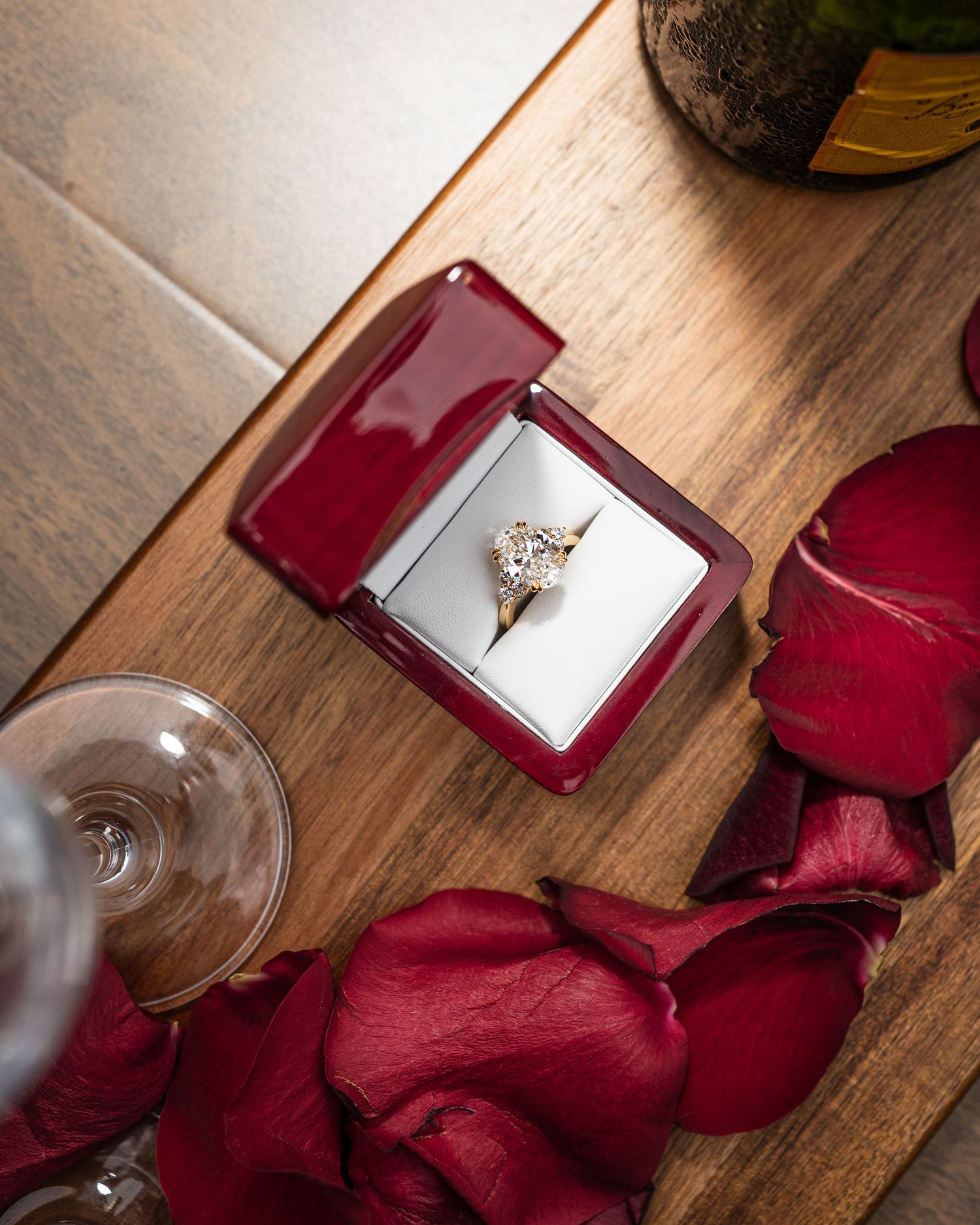 A diamond engagement ring in a red velvet box surrounded by rose petals and a wine glass on a wooden surface.