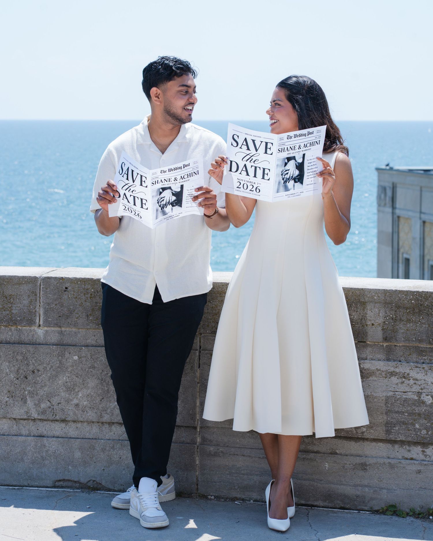 Couple holding Save the Date 2026 signs by the ocean, dressed in white for an engagement photo shoot.