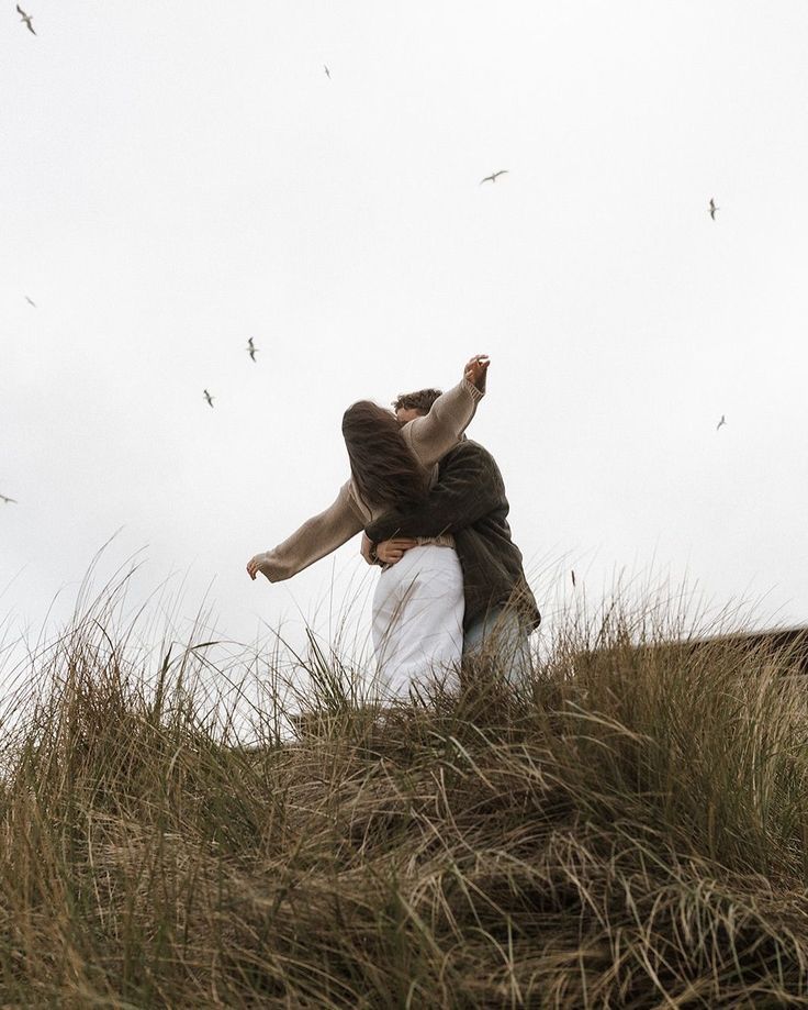 Person dancing freely on grassy dunes with arms outstretched, birds flying overhead against an overcast sky.